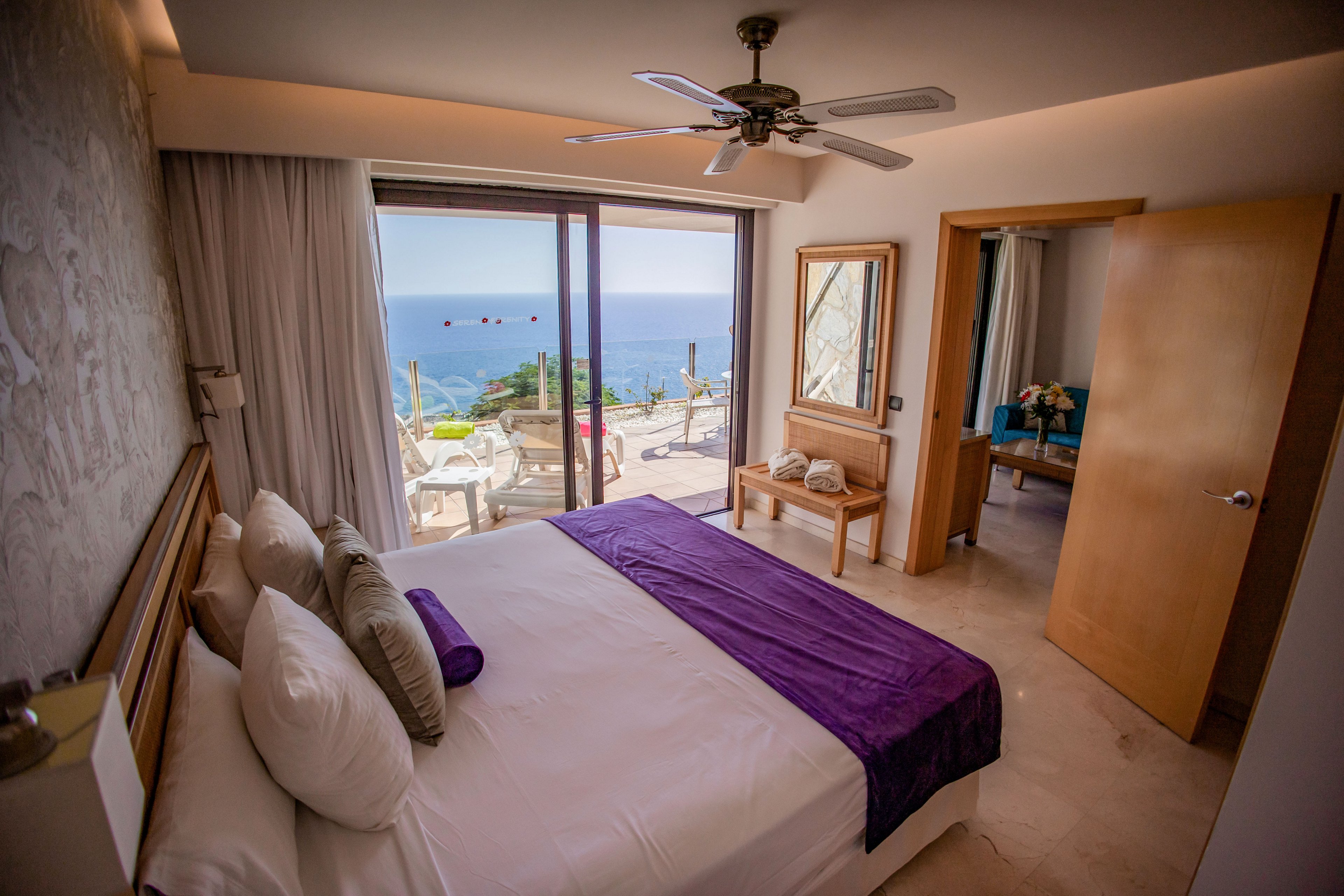 A neatly made resort bedroom features a large bed with white linens and a purple runner facing sliding glass doors. Beyond the doors, a balcony overlooks a bright blue ocean and clear sky. The room includes a ceiling fan, light-colored tile floor, and warm recessed lighting. A wooden bench, mirror, and an open door leading to a sitting area add to the inviting, vacation atmosphere.