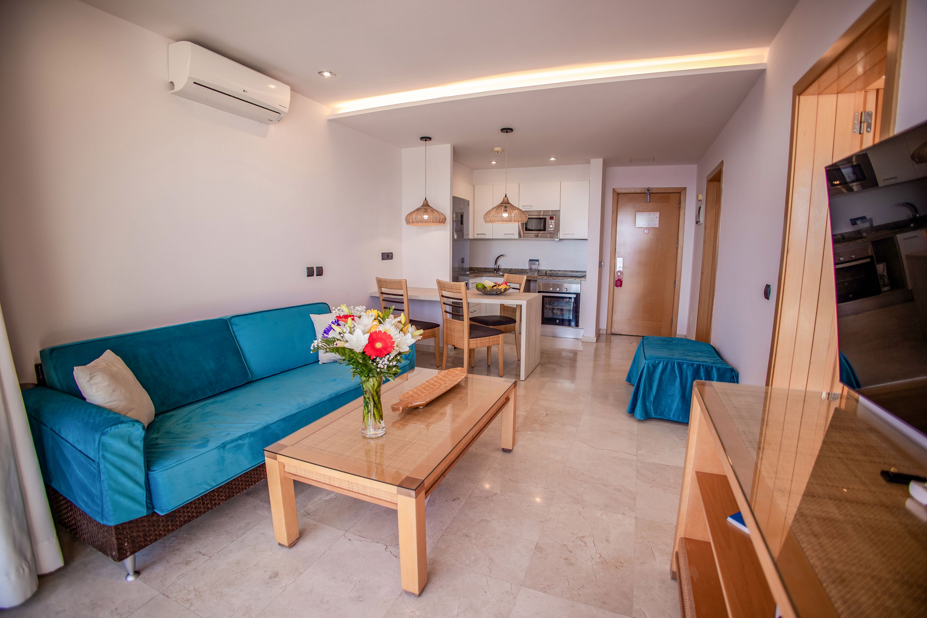 Bright open-plan apartment interior featuring a teal sofa and wooden coffee table with a flower bouquet. The space extends into a compact kitchen area with stainless steel appliances and pendant lights above a dining table. Neutral walls, tiled flooring, and recessed ceiling lighting create a clean, contemporary atmosphere. A wall-mounted air conditioner and doorway in the background suggest a comfortable vacation or rental accommodation.