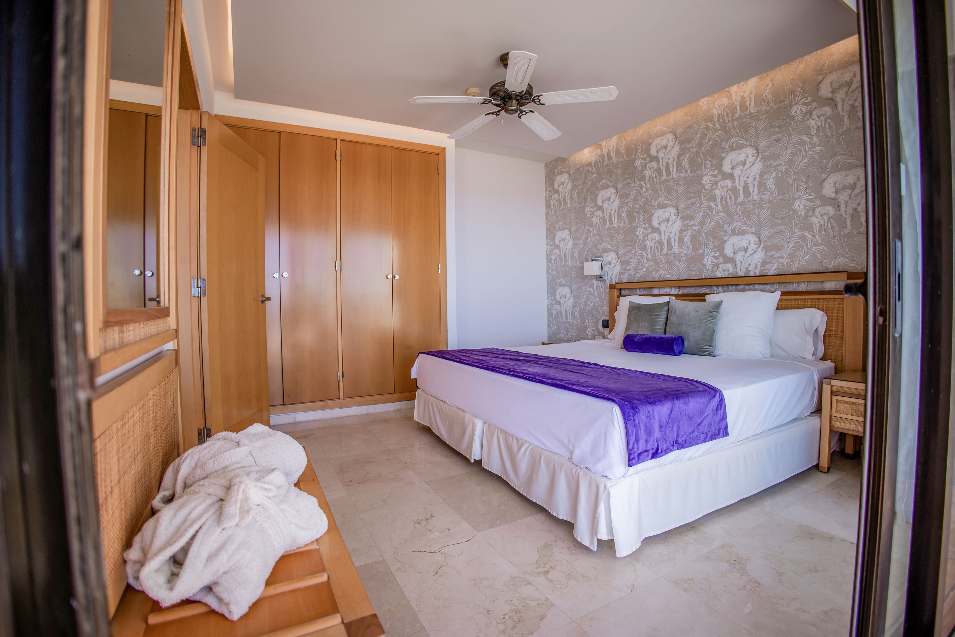A neatly made double bed with white linens and a purple runner is the focal point of this bright hotel bedroom. The room features light wood furniture, including a large wardrobe and bedside tables, and a ceiling fan overhead. A patterned wall with animal illustrations adds decorative interest behind the bed. A folded white robe rests on a bench near the entrance, suggesting a comfortable, relaxing stay.