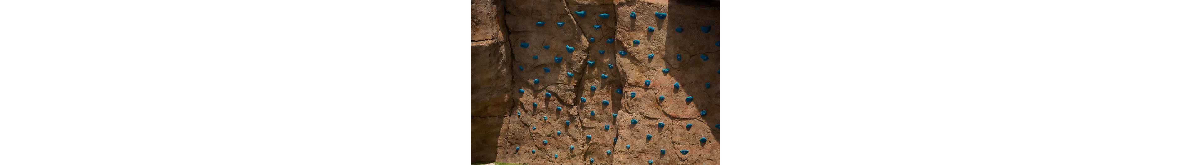 The image shows an artificial rock climbing wall designed to resemble natural stone. Numerous bright blue climbing holds are attached across the vertical surface, creating various routes and difficulty levels. The wall appears outdoors, with sunlight casting shadows that emphasize the textured rock-like panels. No people are present, highlighting the structure and climbing features themselves.