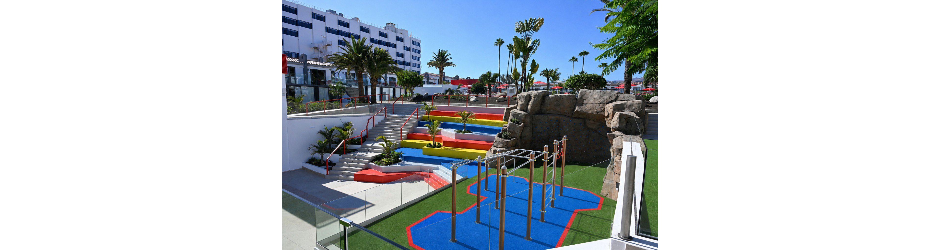 An outdoor playground with bright blue, red, yellow, and green surfaces is set within a modern resort complex. The play area features climbing bars, tiered seating or play platforms, artificial grass, and a large rock structure. Palm trees and landscaped plants surround the space, adding a tropical atmosphere. In the background, a multi-story white building and other resort facilities are visible under a clear blue sky.