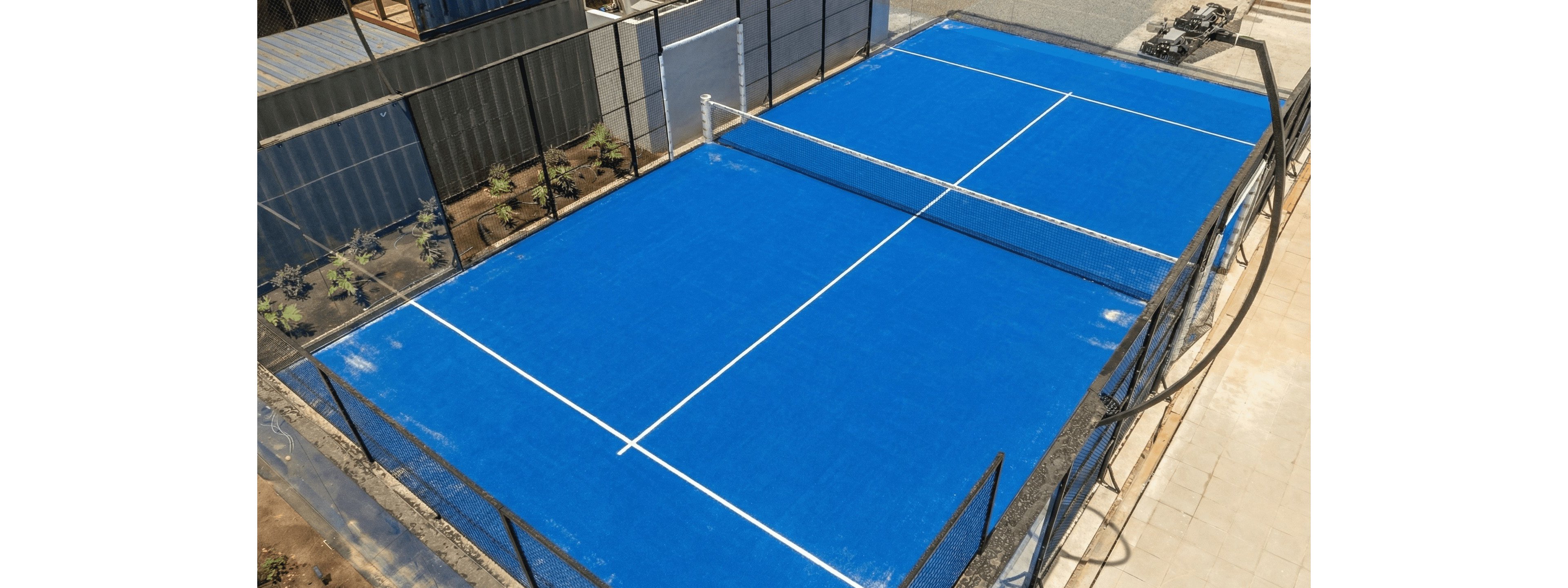 An empty blue tennis court is seen from an elevated perspective in an outdoor setting. White boundary and center lines clearly mark the playing area, with a net running across the middle. The court is surrounded by fencing and nearby paved walkways, with some plants visible along one side. Bright daylight highlights the vivid blue surface and clean geometric layout of the court.