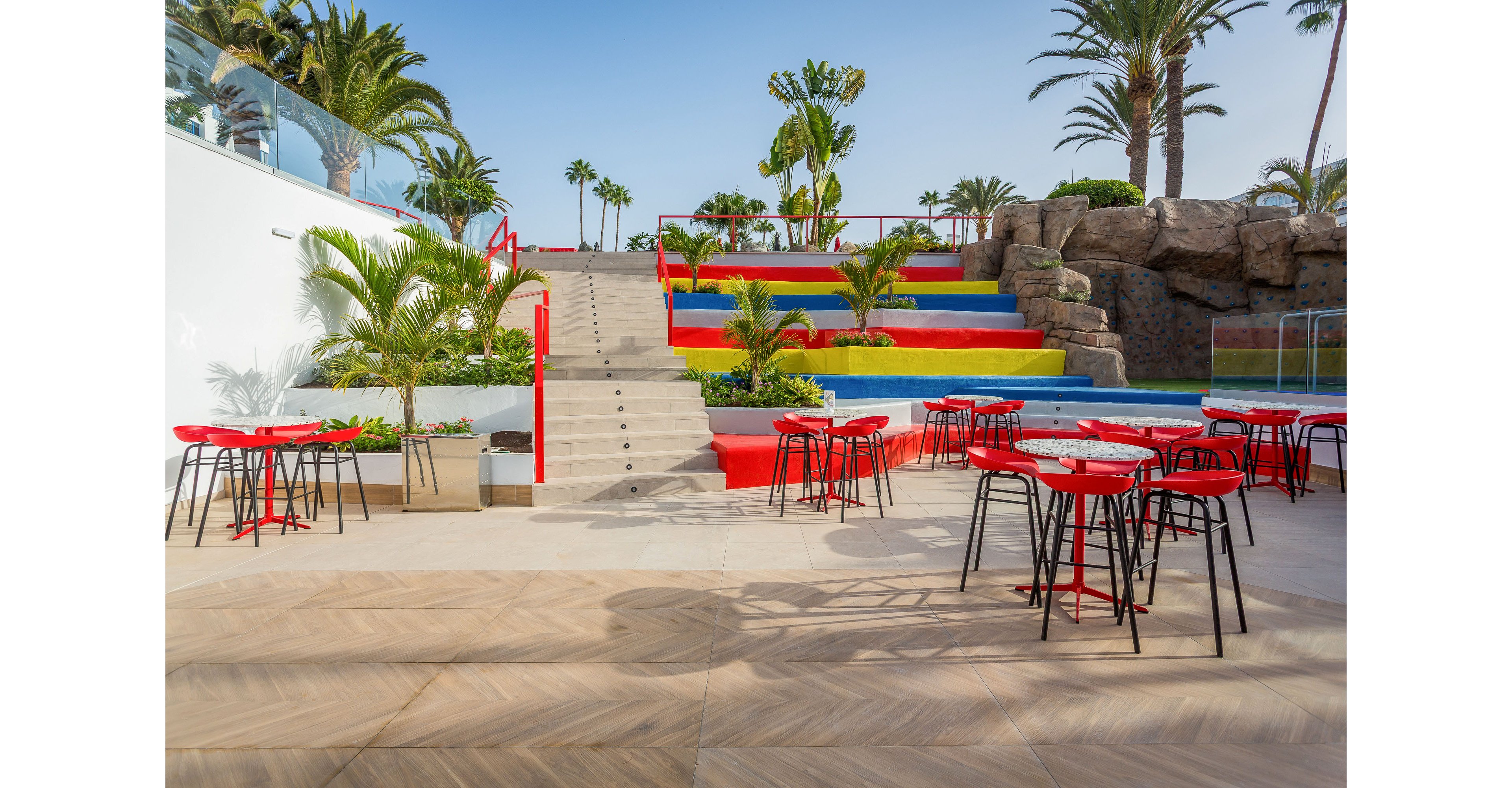 An open-air terrace features red metal tables and high stools arranged on a light tiled floor. A central staircase leads up between vibrant multicolored tiered seating in red, yellow, blue, and green. Palm trees and tropical plants surround the area, adding a lush resort-like atmosphere. The scene is bright and sunny, suggesting a relaxed social or dining space in a warm climate.