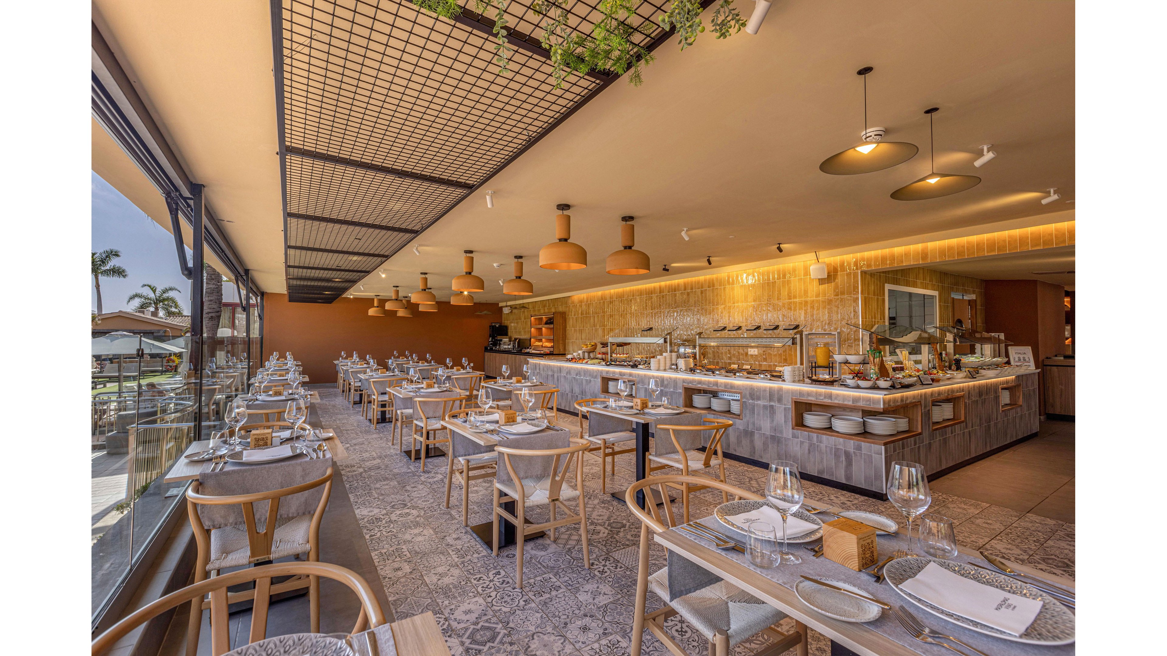 Contemporary restaurant interior featuring a long buffet counter and neatly arranged tables. The space is bright and sunlit, with large floor-to-ceiling windows overlooking an outdoor area. Wooden chairs, patterned floor tiles, and warm pendant lighting create a cozy yet modern atmosphere. Tables are set with plates, cutlery, and wine glasses, ready to welcome guests.