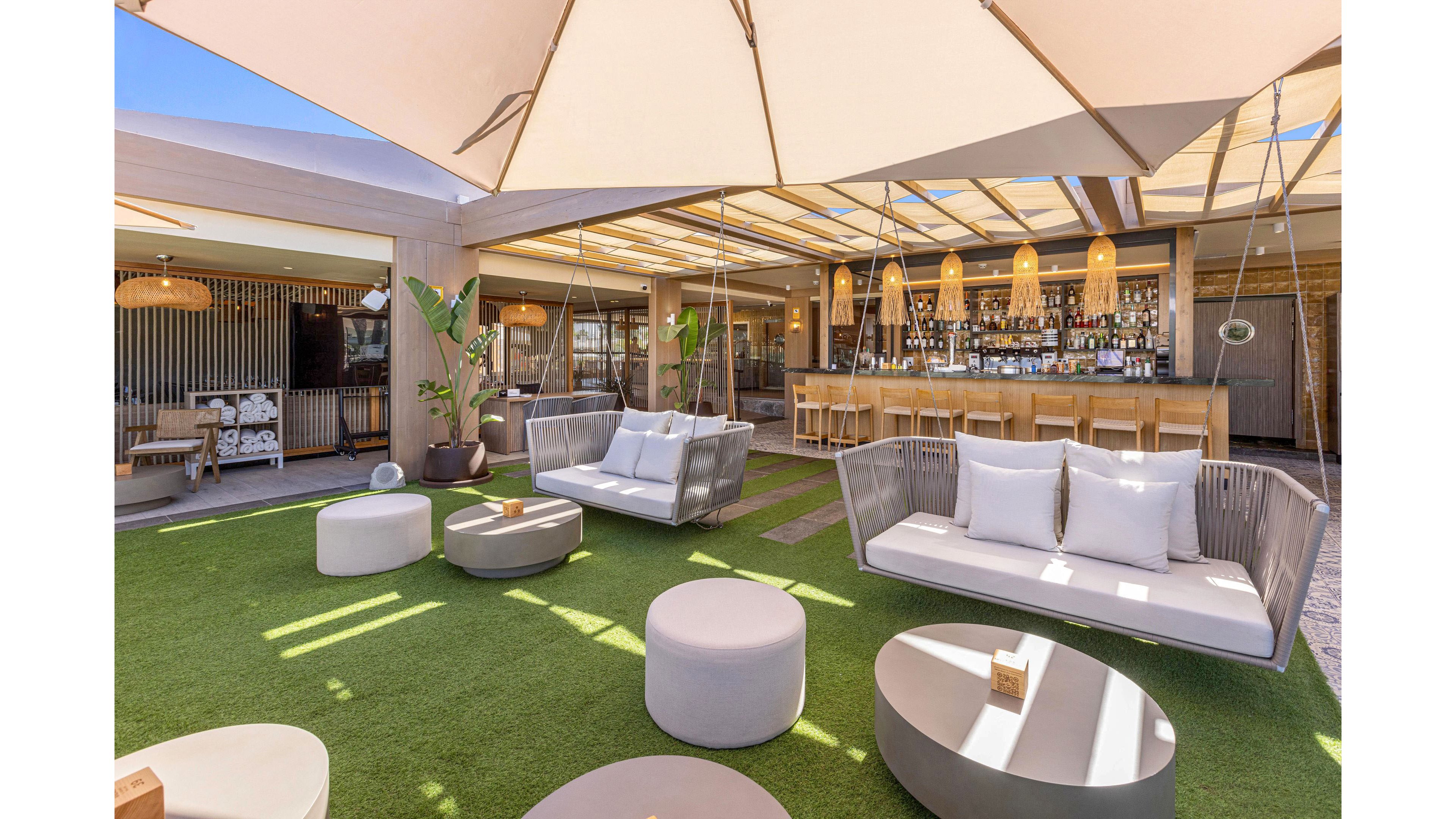 A contemporary outdoor lounge area features cushioned sofas and round ottomans arranged on a green artificial grass floor. A large canopy and pergola roof provide shade while allowing natural light to filter through. In the background, a fully stocked bar with high stools and warm pendant lighting creates a relaxed social atmosphere. Neutral tones, clean lines, and natural textures give the space a modern, resort-like feel.