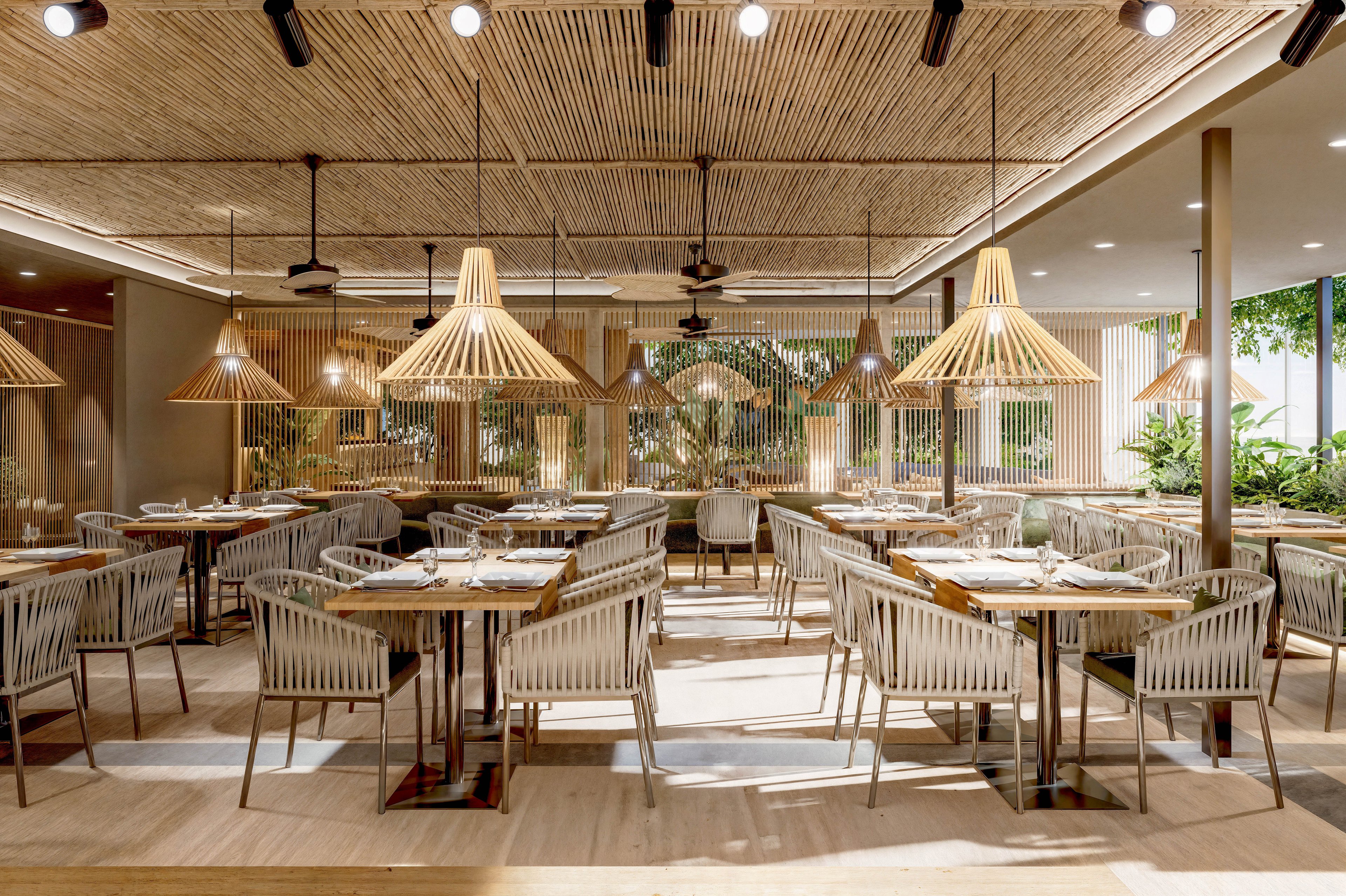 A contemporary restaurant dining area features neatly arranged tables and chairs in a spacious, open layout. The ceiling is lined with wooden slats and distinctive pendant lights that create a warm, tropical ambiance. Neutral tones, natural wood, and woven textures dominate the design, complemented by greenery along the perimeter. The setting appears calm and inviting, ideal for relaxed dining or social gatherings.