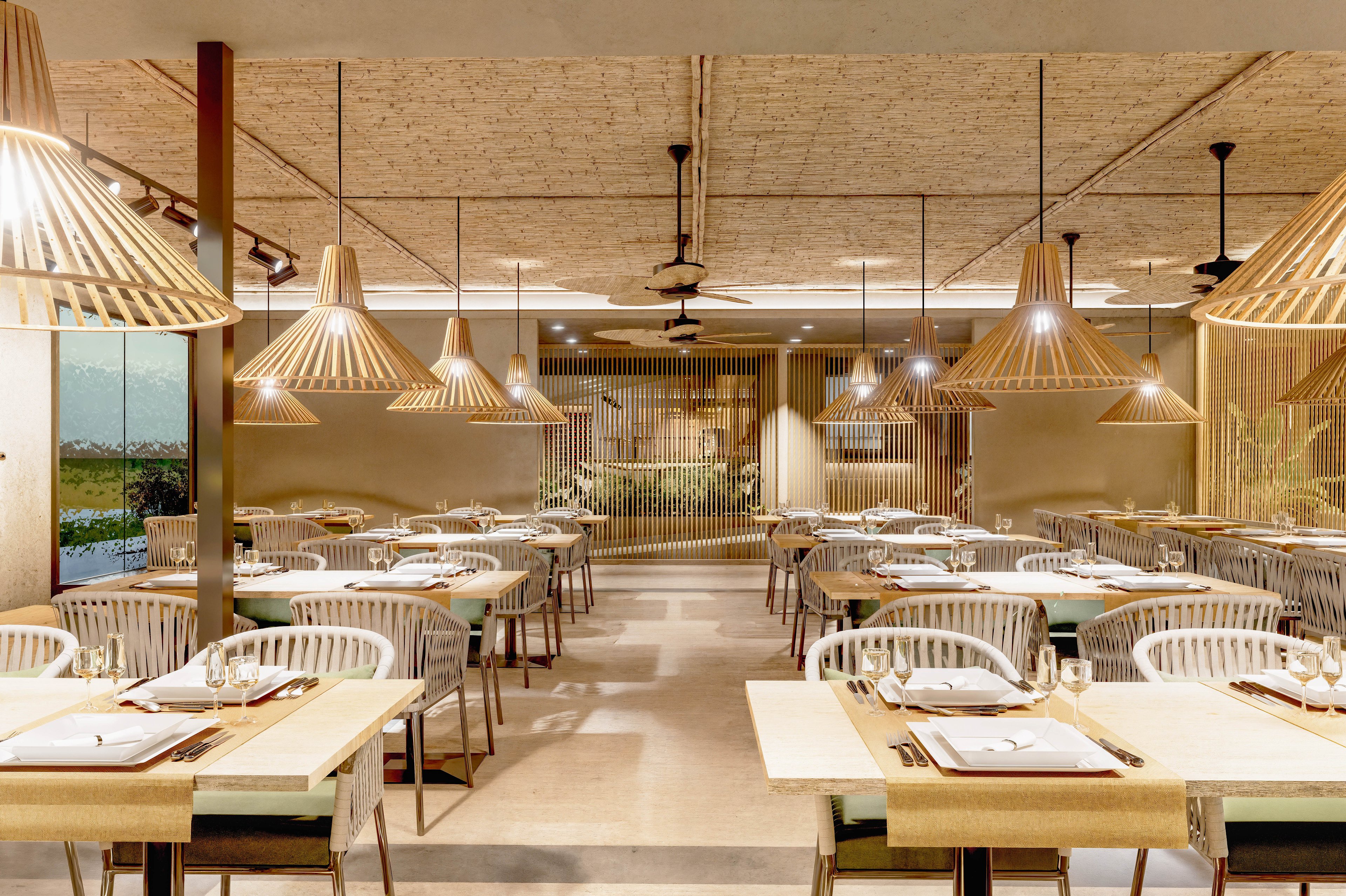 A contemporary restaurant dining area features neatly arranged tables and chairs in a warm, neutral palette. Wooden pendant lights hang from the ceiling, casting soft illumination over the place settings and creating a cozy atmosphere. The space is open and symmetrical, with light wood accents, slatted partitions, and clean architectural lines. No people are present, emphasizing the calm, orderly design and inviting ambiance.