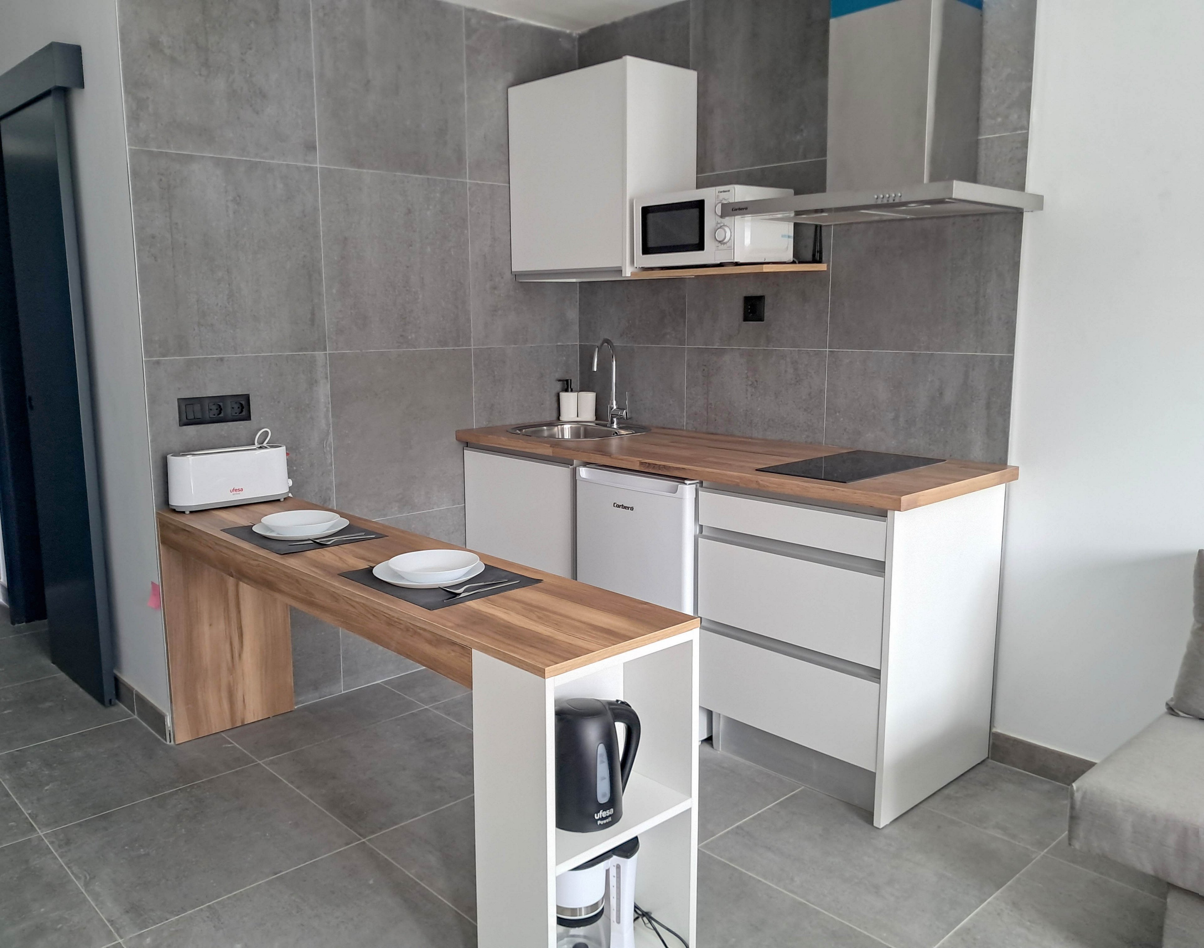 A modern compact kitchenette features white cabinets and a wooden countertop against large gray tiled walls. The space includes a small sink, electric cooktop, microwave, and under-counter refrigerator. A narrow breakfast bar with two place settings extends from the counter, creating a functional dining area. A kettle and toaster add everyday practicality to the clean, minimalist design.
