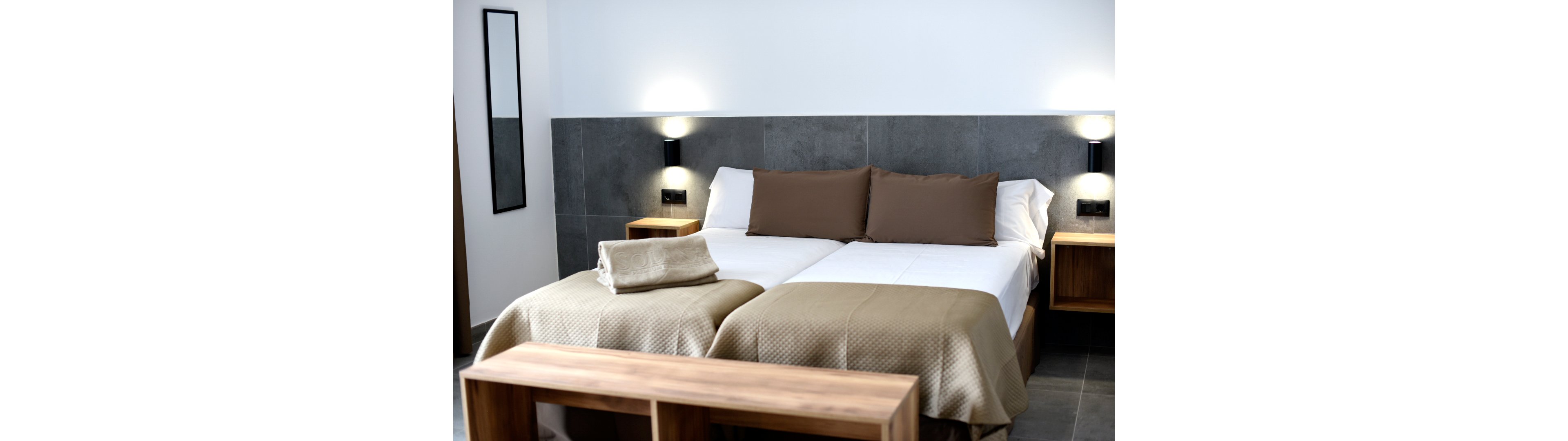 A contemporary twin bedroom features two single beds pushed together, topped with beige quilts and brown accent pillows. Freshly folded towels are placed neatly at the foot of one bed, suggesting a hotel or guest accommodation. The room has a minimalist design with dark gray wall tiles, wooden bedside tables, and a matching wooden bench. Wall-mounted lights and a tall mirror add to the clean, modern aesthetic.