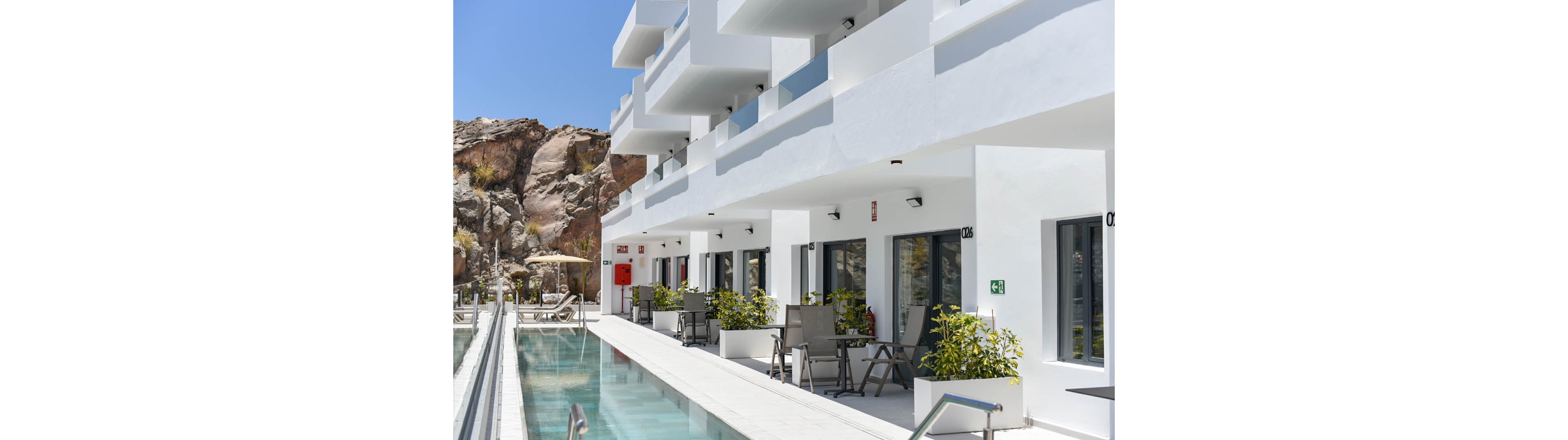 A modern white hotel building features ground-floor rooms opening onto private terraces beside a long, narrow swimming pool. Lounge chairs and small tables line the poolside, creating a relaxed vacation atmosphere under clear blue skies. Rocky cliffs rise behind the structure, contrasting with the clean minimalist architecture. The bright sunlight emphasizes the crisp white walls and reflective water surface.