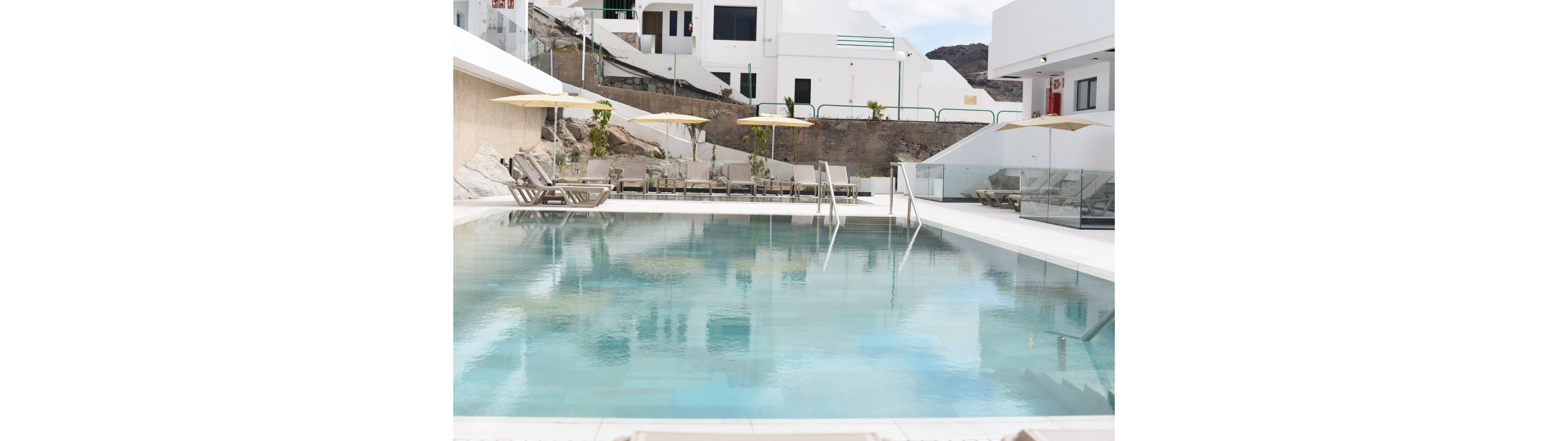 A clear, rectangular swimming pool sits at the center of a modern hotel or apartment complex. Beige sun loungers and yellow umbrellas line the far side of the pool, creating a relaxed resort atmosphere. White contemporary buildings and glass railings surround the pool area, with rocky hills visible in the background. The scene appears calm and quiet, suggesting an off-peak or early day setting.