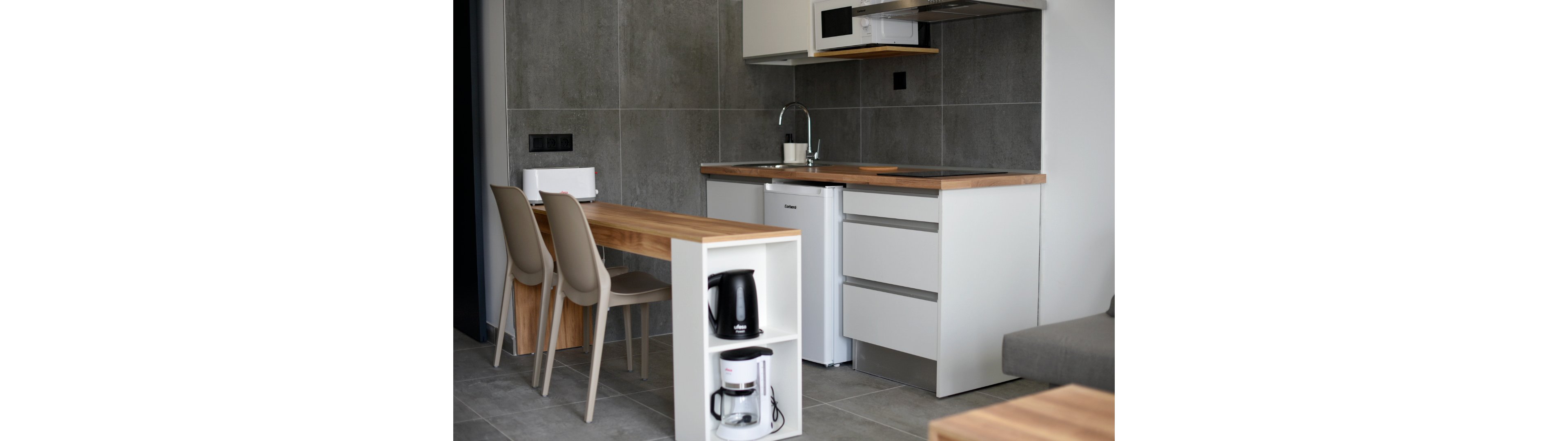 A modern compact kitchenette features a wooden countertop and minimalist white cabinetry. A small dining table with two beige chairs extends from the counter, creating a combined cooking and eating area. Built-in shelves hold a black electric kettle and a white coffee maker, while a mini fridge and microwave are integrated into the design. Grey tiled walls and floors give the space a clean, contemporary look.