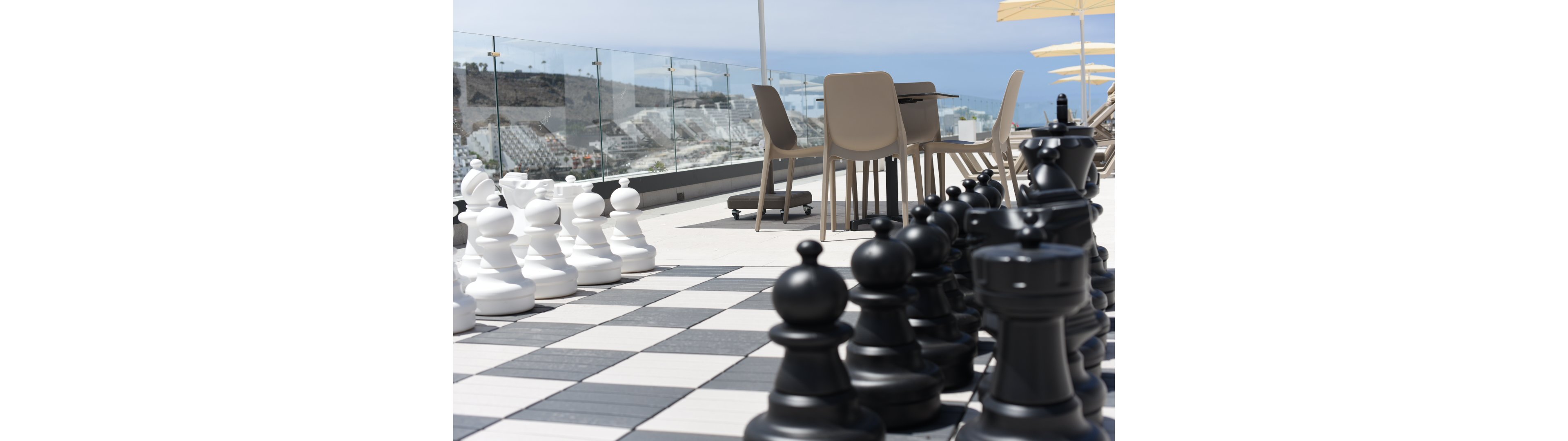 A large outdoor chessboard with oversized black and white pieces is set up on a modern rooftop terrace. In the background, a table and chairs sit near a glass railing that reveals a coastal city and ocean view. The bright daylight and clear sky create a relaxed, leisurely atmosphere. The scene combines strategic play with a contemporary, resort-like setting.