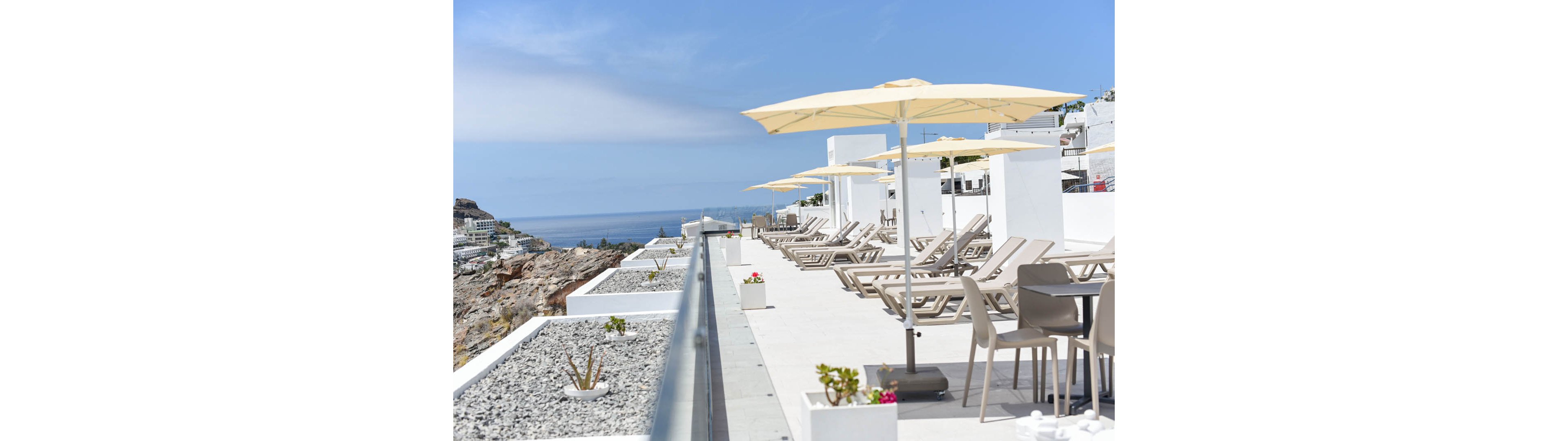 A modern rooftop terrace features rows of beige sun loungers under large cream umbrellas. The space is designed with clean white architecture and minimalist planters along a glass railing. In the distance, a rocky hillside descends toward the sea under a clear blue sky. The scene suggests a tranquil resort environment ideal for sunbathing and relaxation.
