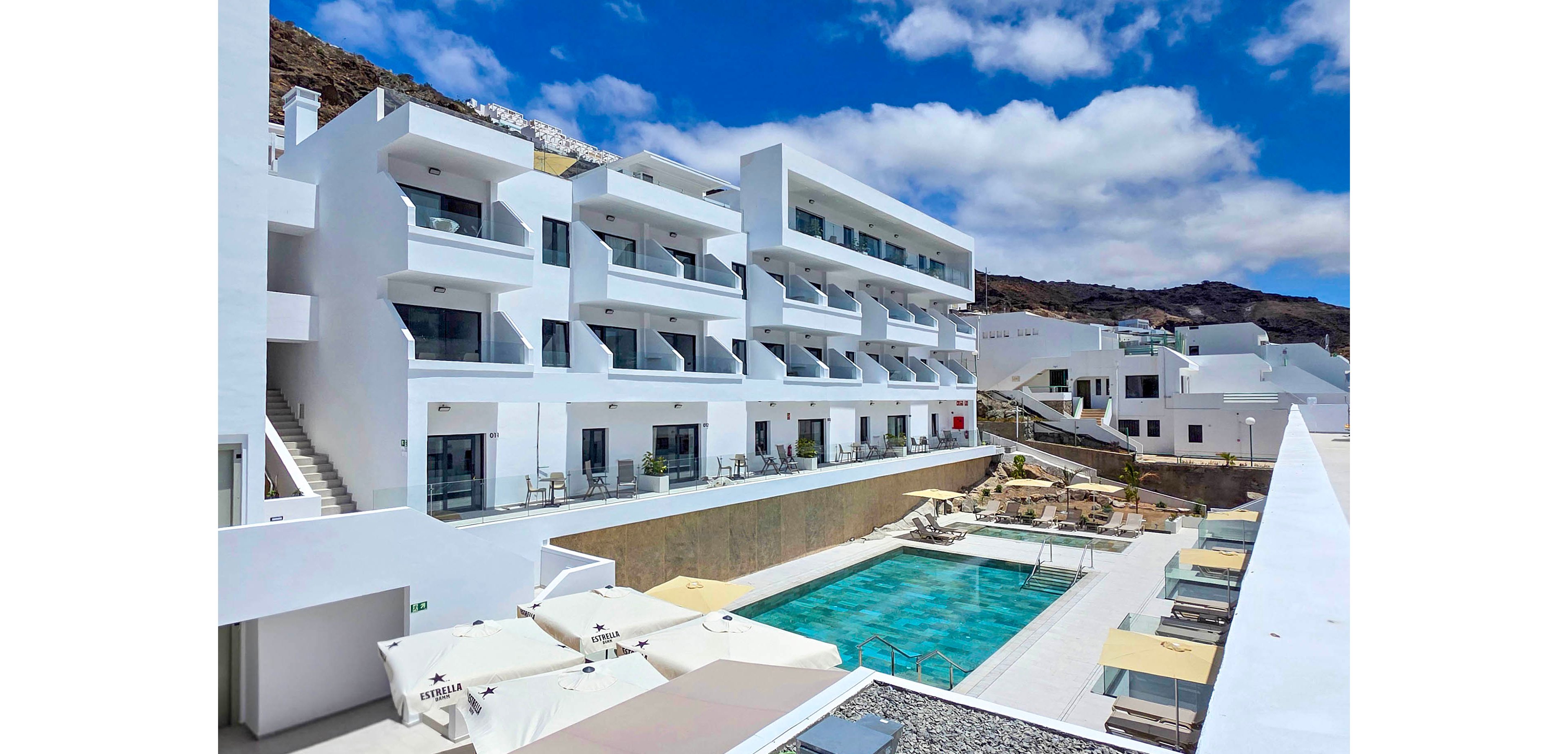 A contemporary white multi-story hotel complex is built into a rocky hillside under a partly cloudy blue sky. The building features rows of balconies overlooking a central outdoor swimming pool and sun terrace. Lounge chairs and umbrellas are arranged neatly around the pool, suggesting a relaxing vacation setting. Additional white structures and terraces extend into the background, emphasizing the resort’s clean, minimalist architecture.