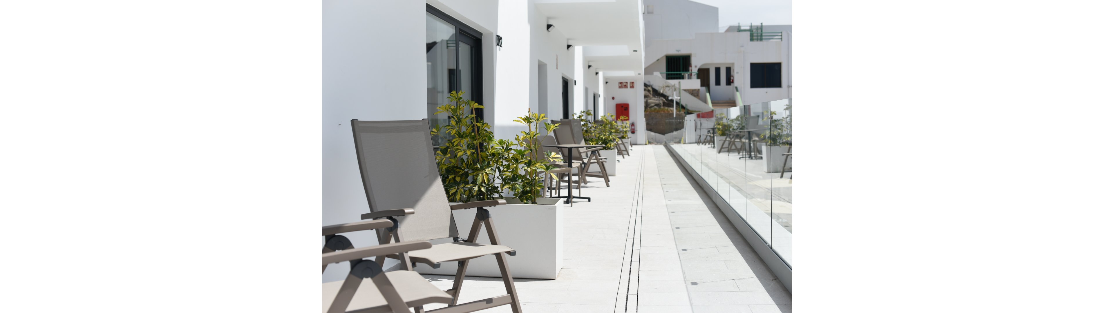 A row of beige outdoor chairs lines a modern white terrace. Potted green plants sit between the chairs, adding color to the minimalist architecture. Glass railings on one side reflect the bright sunlight and clean lines of the building. The scene appears calm and quiet, suggesting a residential or vacation apartment complex.