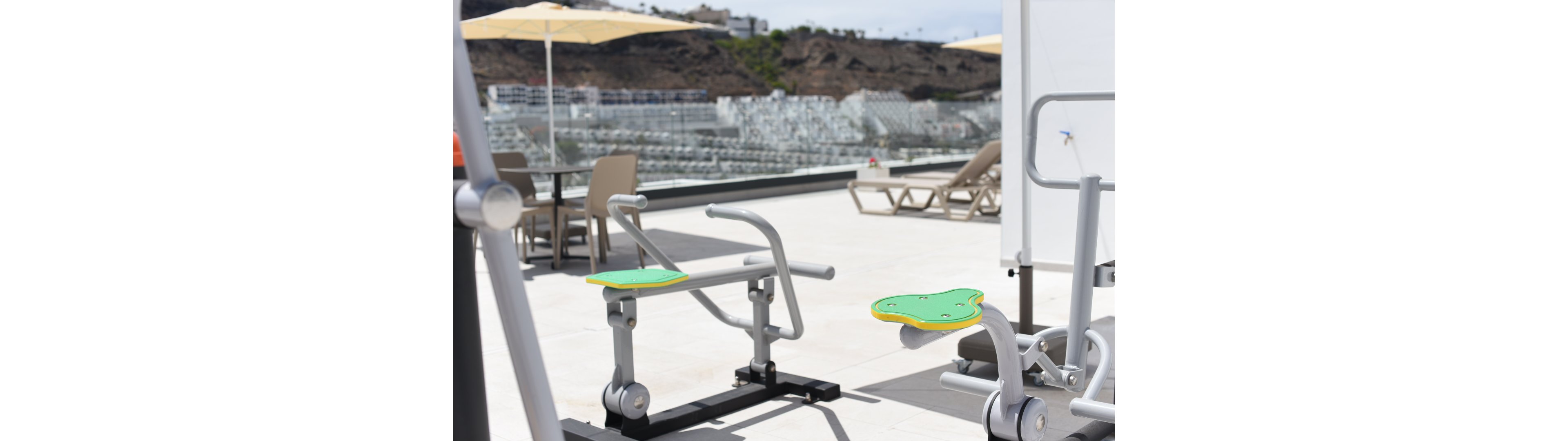 An outdoor fitness station with metal exercise machines is set up on a bright rooftop terrace. The equipment features green and yellow seats and handles, positioned on a smooth tiled surface. In the background, lounge chairs, tables with umbrellas, and a hillside town or resort are visible. The scene is sunlit and relaxed, combining leisure seating with workout facilities.