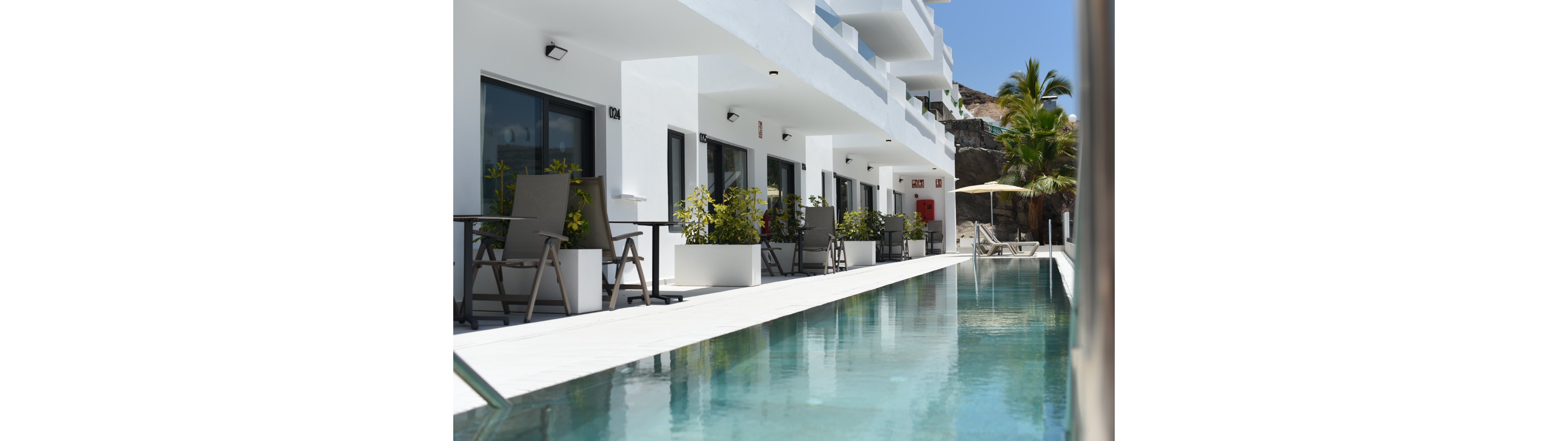 A sleek white resort building lines a narrow outdoor swimming pool. Private terraces with chairs, tables, and potted plants face the water, creating an inviting relaxation area. The scene is bright and sunny, with reflections shimmering on the clear blue pool surface. Palm trees and rocky hills in the background suggest a warm, tropical or coastal destination.