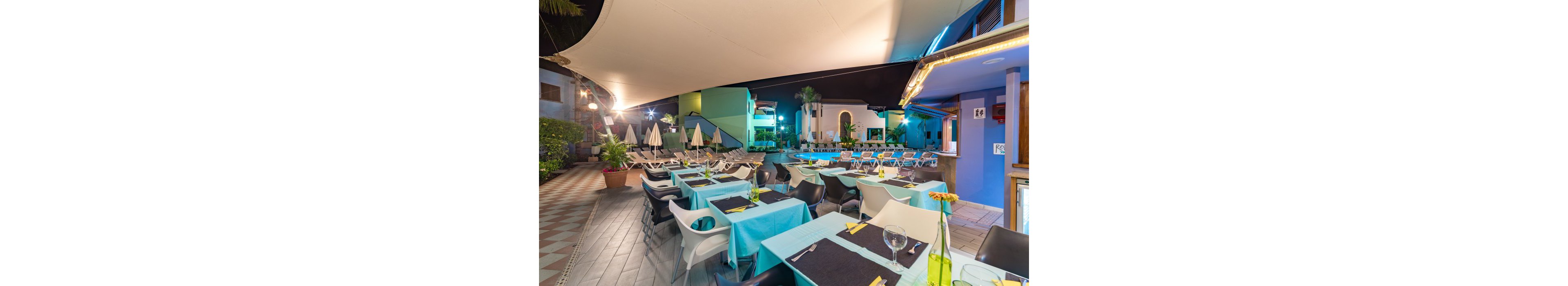 An outdoor restaurant terrace is set up beside a swimming pool at night. Tables are neatly arranged with turquoise tablecloths, black runners, and glassware under a large canopy. Sun loungers and closed umbrellas line the pool area in the background, suggesting a resort or hotel setting. Colorful building facades and warm lighting create a relaxed, vacation atmosphere.