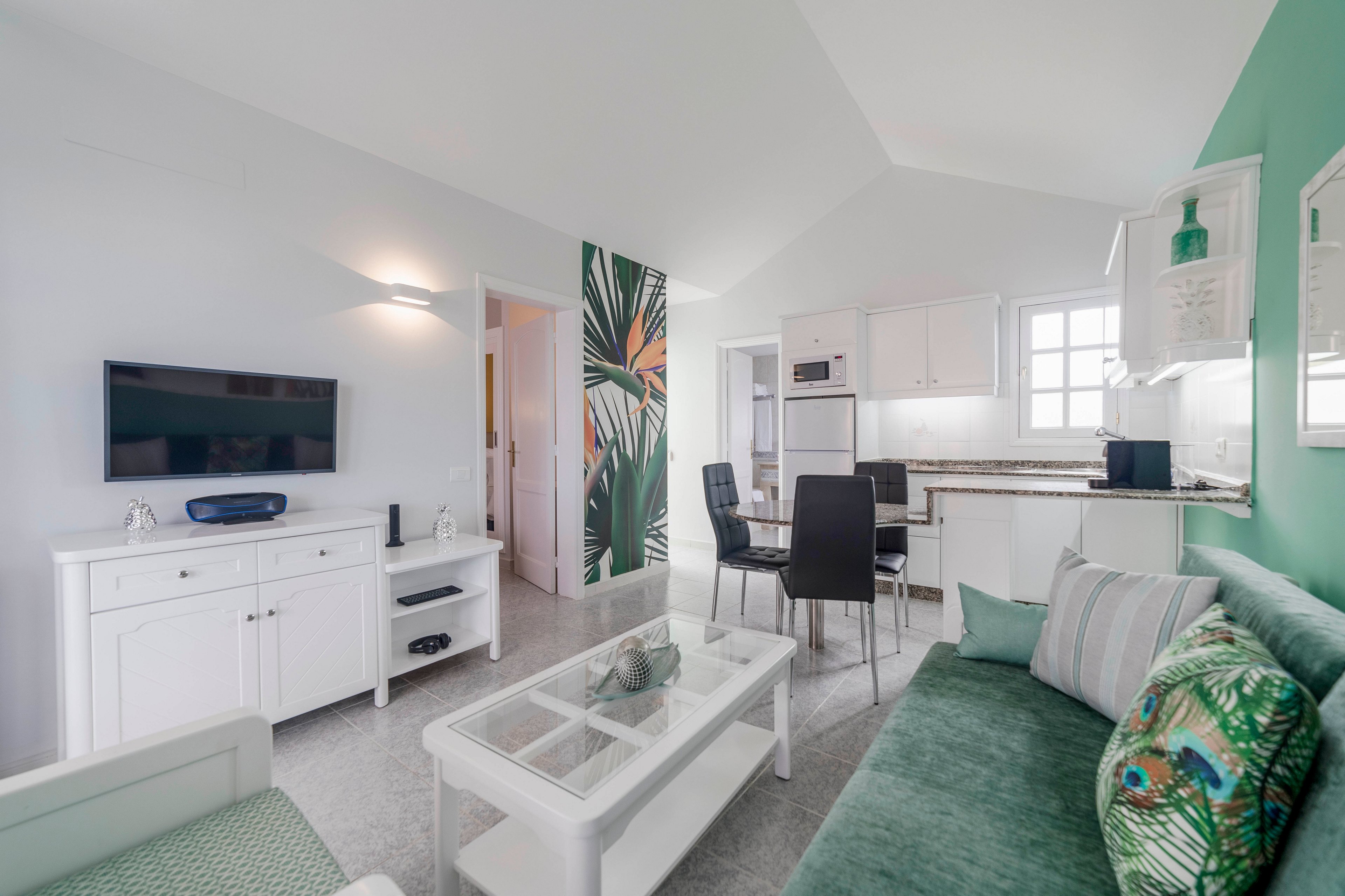 Contemporary open-plan living room with a green sofa and glass coffee table leads into a compact kitchen and dining area. White cabinetry, built-in oven, and black dining chairs create a clean, minimalist look. A bold tropical leaf wall mural and green accent wall add color and visual interest. The space includes a wall-mounted flat-screen TV, decorative cushions, and soft natural light from a nearby window.