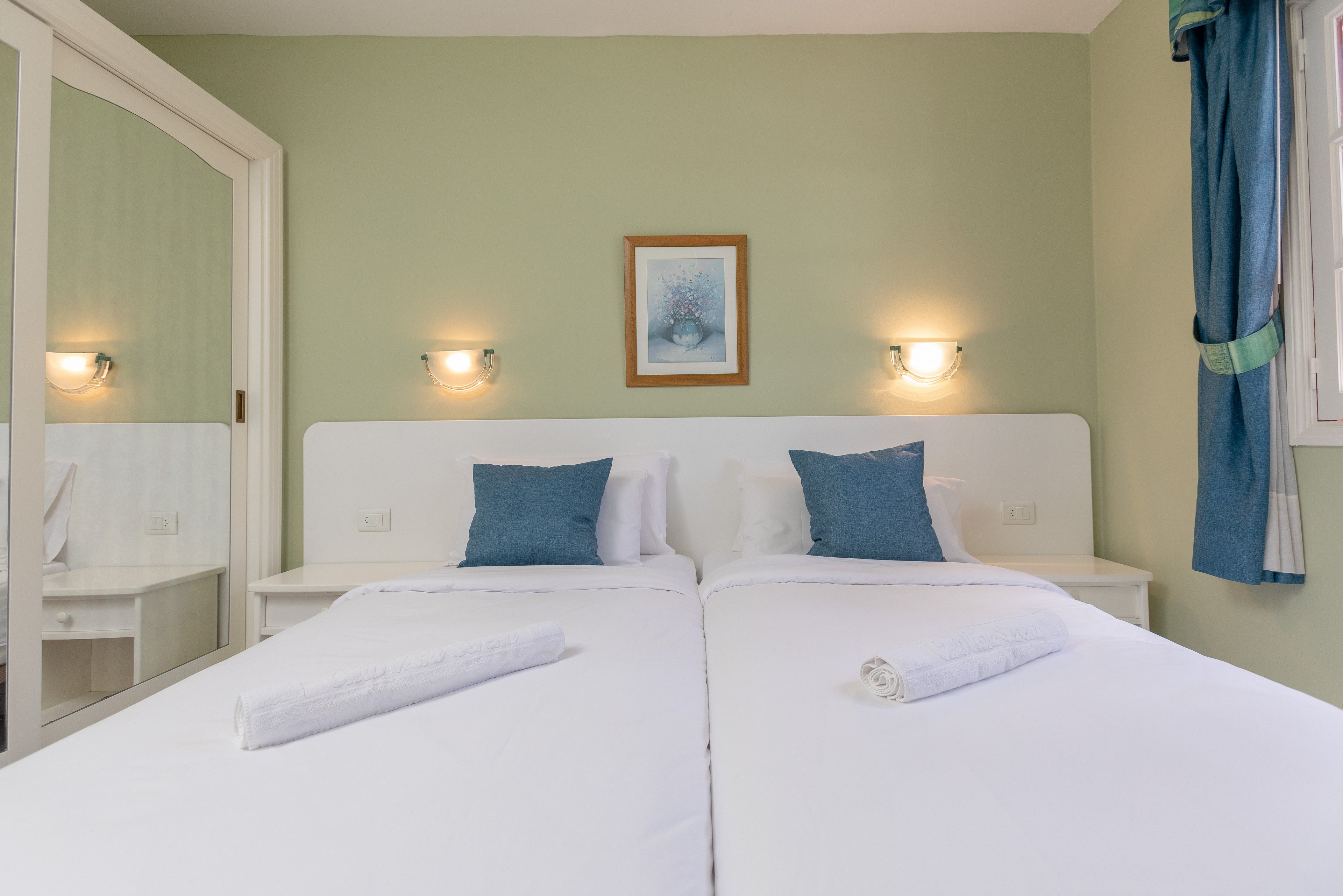A neatly arranged twin bedroom features two single beds pushed together with crisp white linens. Rolled white towels are placed at the foot of each bed, and blue accent pillows add a touch of color. The room has light green walls, a framed artwork above the headboard, and wall-mounted lights providing soft illumination. A mirrored wardrobe and a window with blue curtains enhance the sense of space and natural light.
