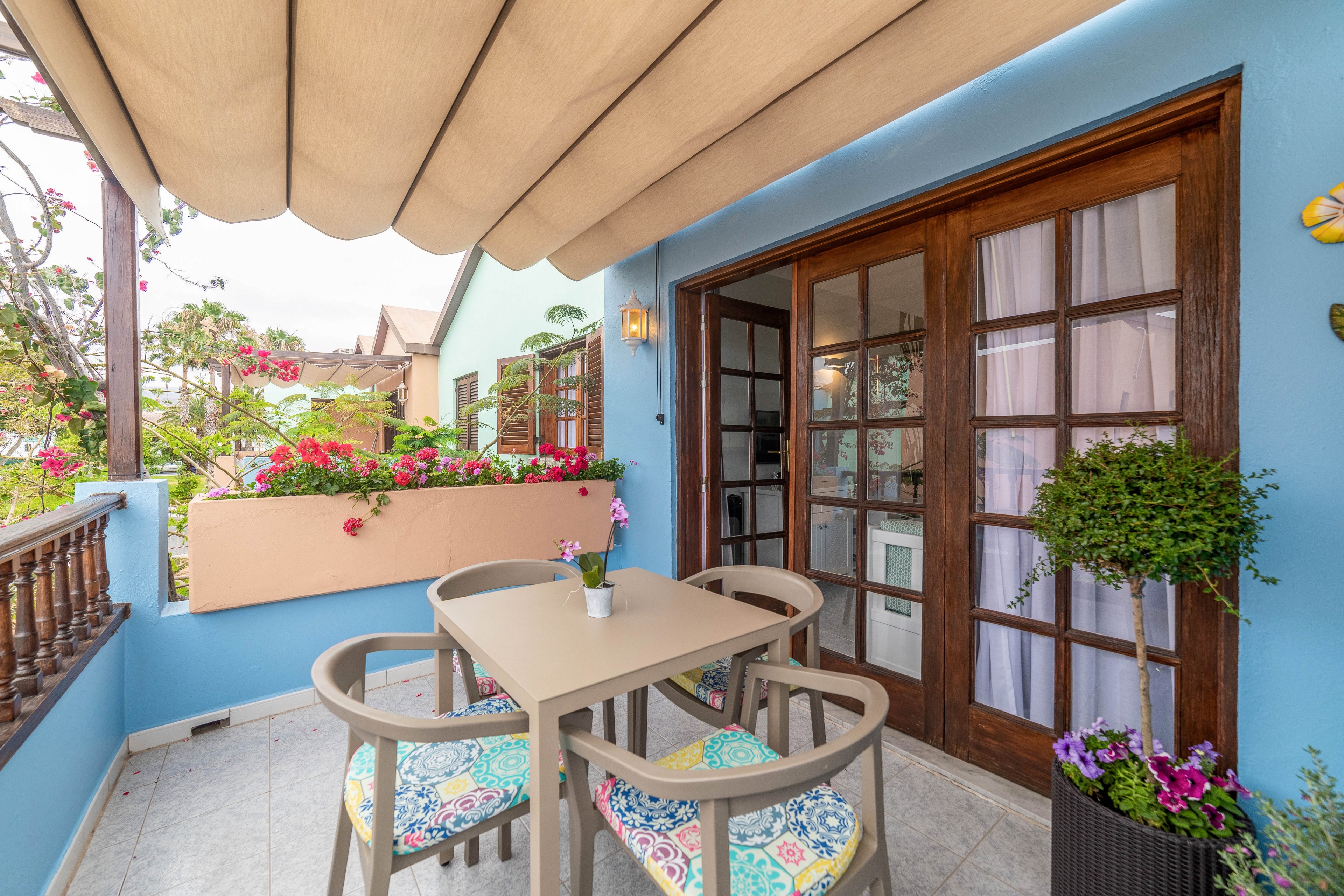 A small balcony terrace features a square table with four chairs, each with vibrant patterned cushions. The space is attached to a home with light blue walls and wooden-framed glass doors covered by sheer curtains. A beige retractable awning shades the area, while potted plants and a flower box filled with blooming flowers add greenery and color. The setting feels inviting and relaxed, ideal for outdoor dining or casual conversation.