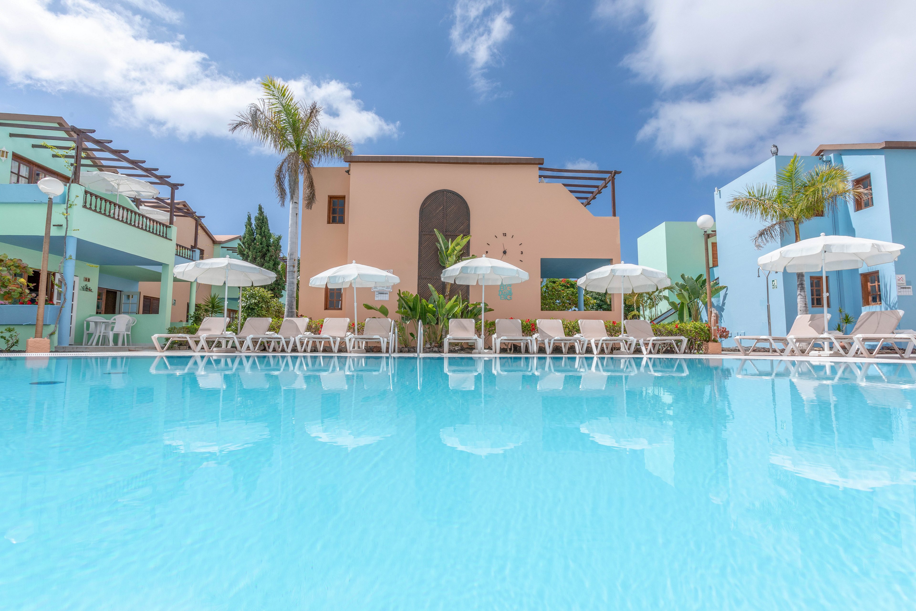 A large, clear blue swimming pool reflects pastel-colored resort buildings under a bright sky. White sun loungers and umbrellas line the poolside, suggesting a relaxing vacation setting. Palm trees add a tropical touch between the modern, geometric structures. The scene is calm and inviting, with no people visible, emphasizing tranquility and leisure.