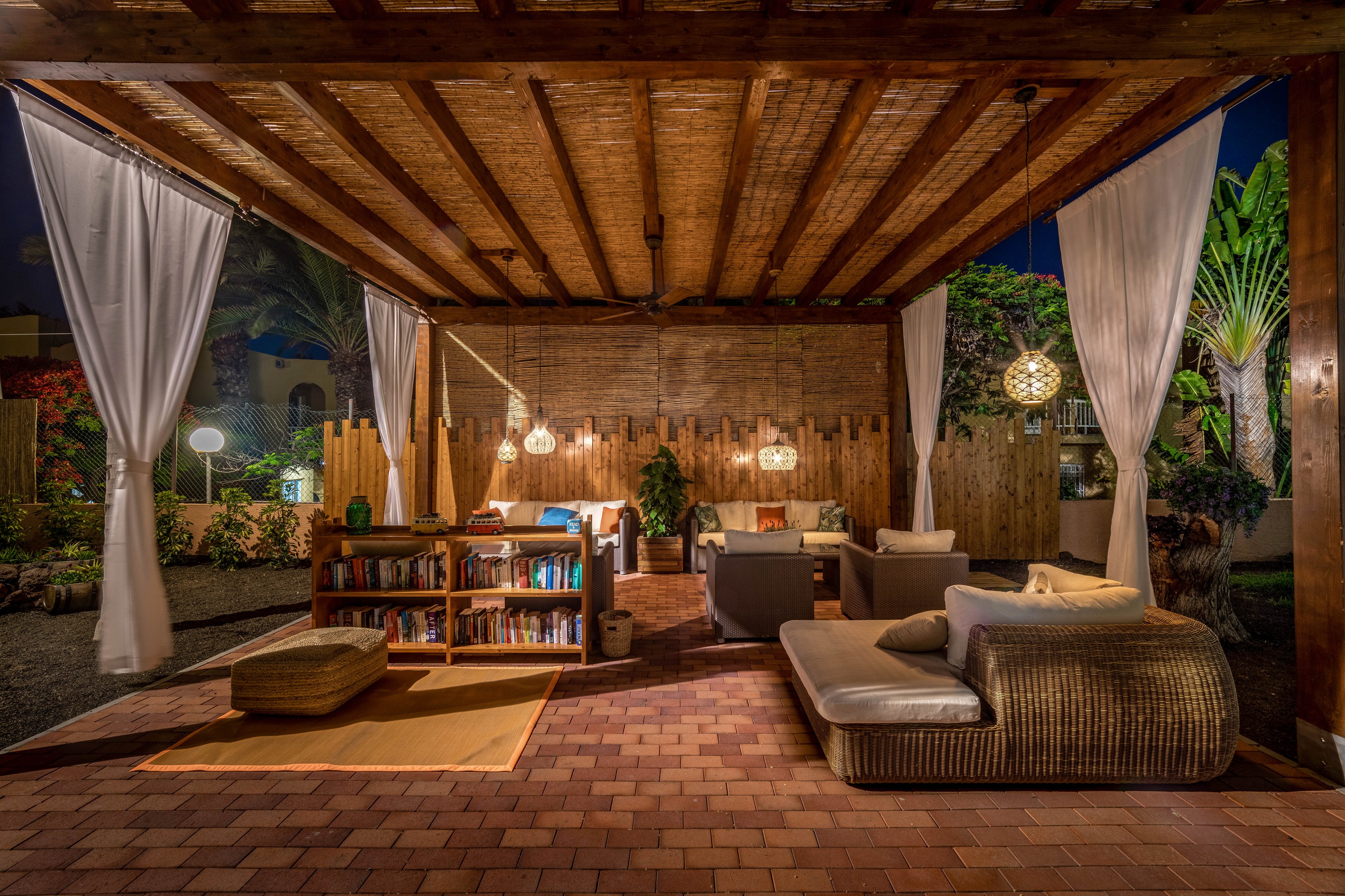 An inviting outdoor lounge area is arranged beneath a wooden pergola at night. Comfortable wicker sofas, cushions, and a low daybed create a relaxed seating space. Hanging pendant lights and lanterns cast a warm glow over a bookshelf and small table. White curtains frame the open sides, while lush tropical plants and trees surround the cozy retreat.