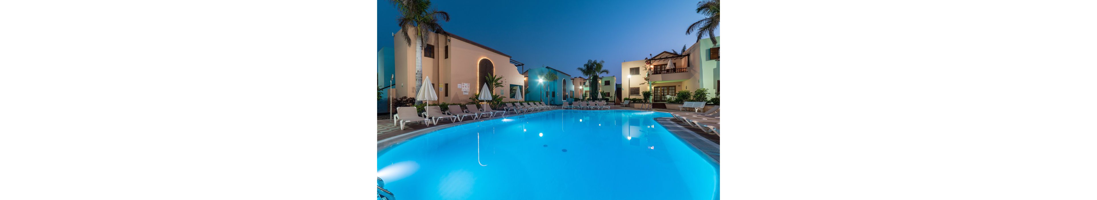 A large illuminated swimming pool sits at the center of a resort courtyard at dusk. Colorful low-rise buildings and balconies surround the pool, with palm trees adding a tropical feel. Numerous empty sun loungers and closed umbrellas line the poolside, suggesting a quiet evening atmosphere. The clear blue water reflects the buildings and deepening twilight sky, creating a calm and inviting vacation scene.