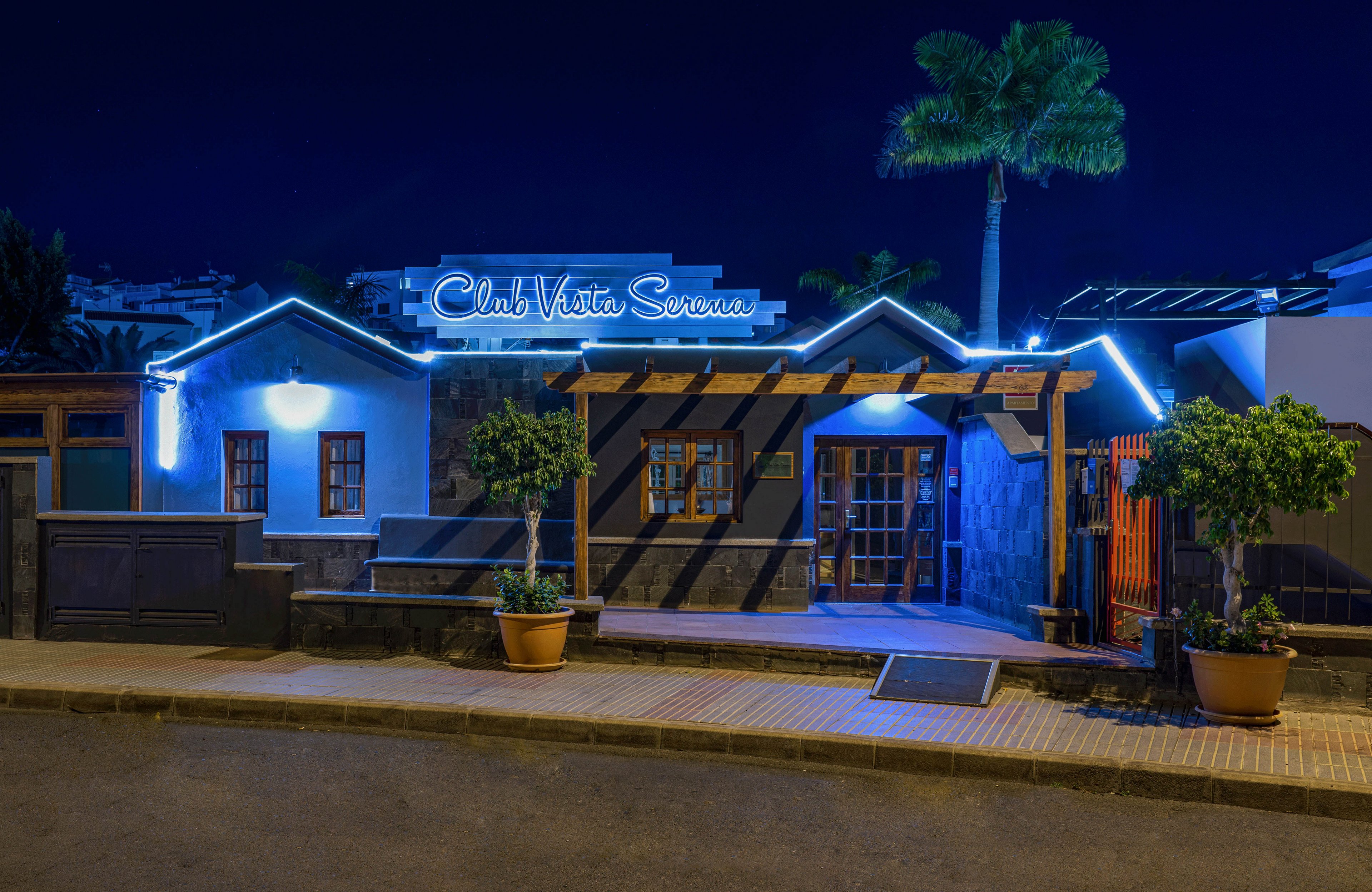 A small club building glows with blue neon lights reading "Club Vista Serena" against the night sky. The structure features white and dark stone walls, wooden doors, and a pergola over the entrance. Potted trees and a palm tree add greenery along the quiet, empty street. Warm interior lighting contrasts with the cool blue exterior illumination, creating a relaxed nightlife atmosphere.