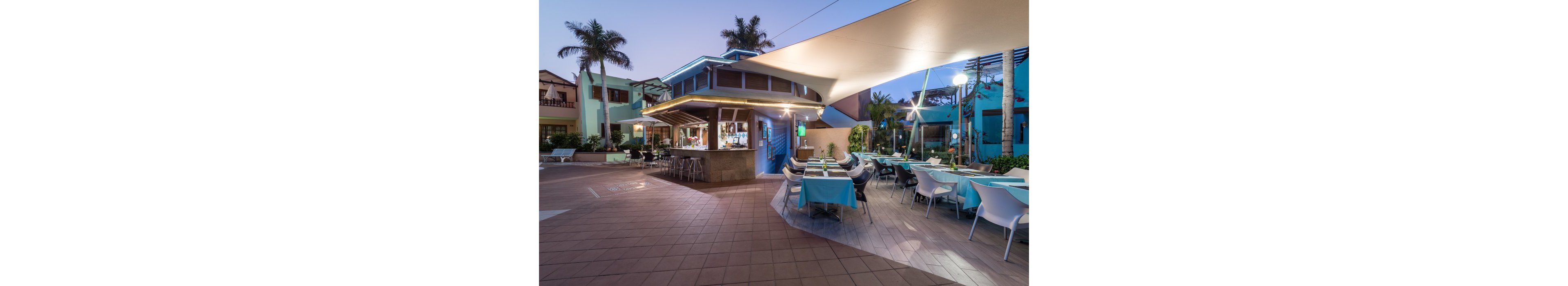 An open-air bar and restaurant is set within a tropical resort courtyard at dusk. Tables with blue and white tablecloths are neatly arranged under a large shade sail canopy. The bar area is illuminated with warm lights, and modern chairs line the counter. Palm trees and colorful resort buildings frame the relaxed, vacation atmosphere.