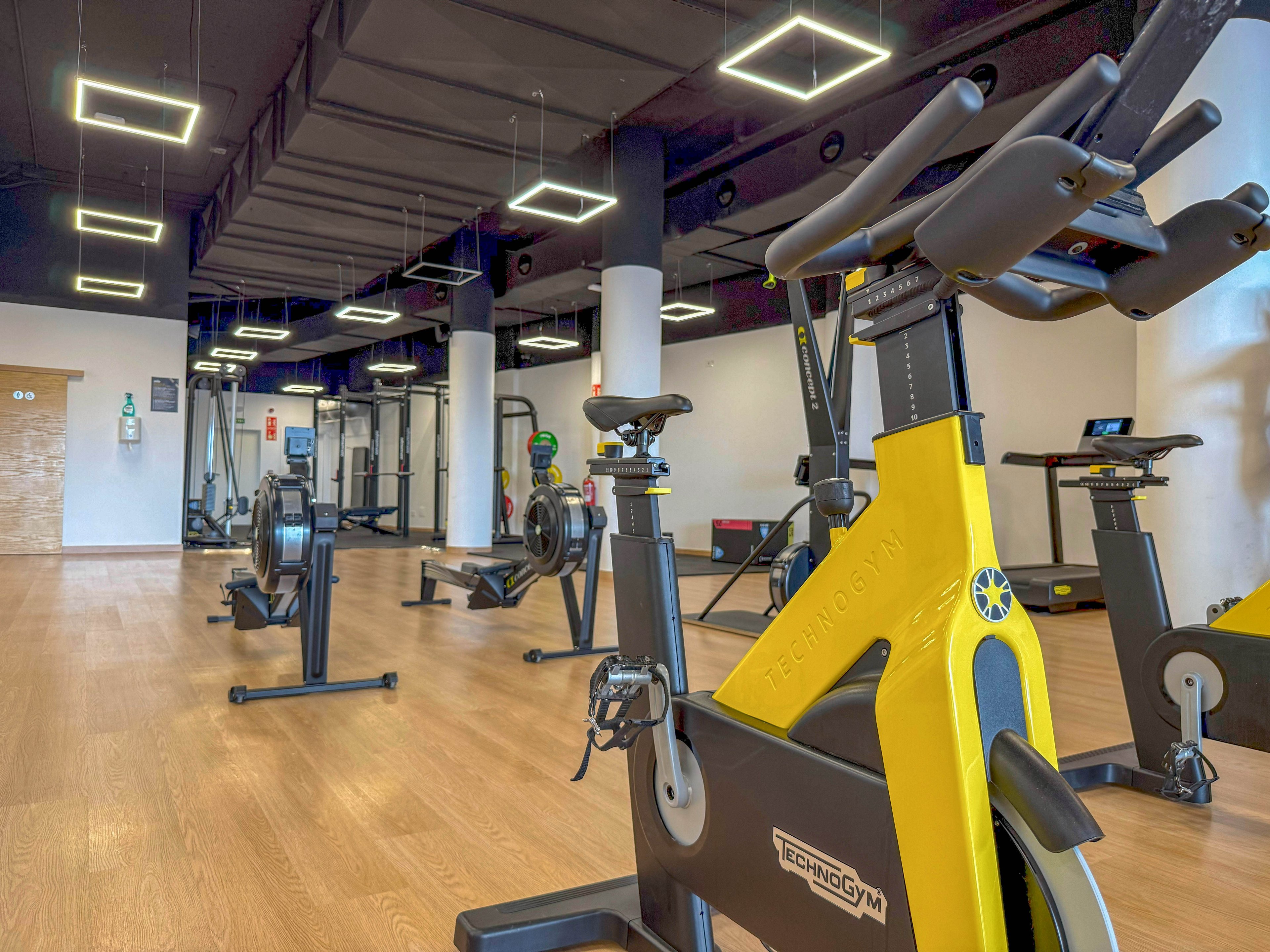 A spacious, well-lit gym features Technogym stationary bikes prominently in the foreground. The setting includes additional fitness equipment such as rowing machines and weight stations, all arranged on a light wooden floor. The ceiling has geometric square lights, and the overall palette is clean and modern with yellow and black accents. No people are present, emphasizing the equipment and open space. The Technogym logo is clearly visible on the exercise bike.