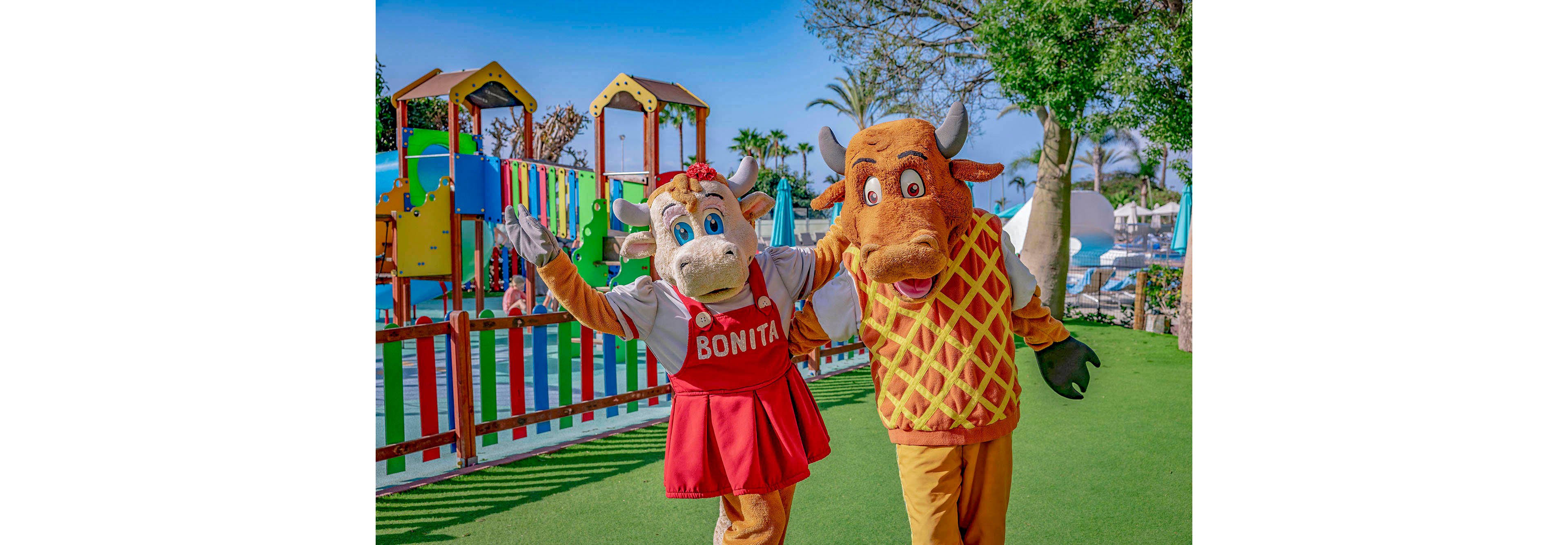 Two people dressed in cheerful cow and bull costumes pose together in front of a vibrant playground. The cow costume features a red dress with the name 'BONITA' clearly visible on the front. The setting is sunny with green artificial grass, colorful play structures, and palm trees in the background. The overall mood is playful and family-friendly.