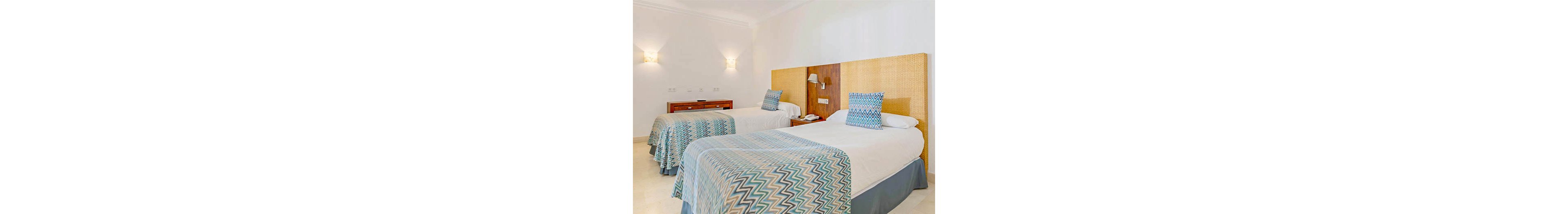 A bright, contemporary hotel room features two twin beds with matching chevron-patterned bedspreads and pillows. The room is decorated in neutral tones with a woven headboard and minimalist wall sconces providing soft lighting. A wooden nightstand with a telephone and a dresser are visible in the background. The overall atmosphere is clean, inviting, and modern. No visible text or numbers are present in the image.
