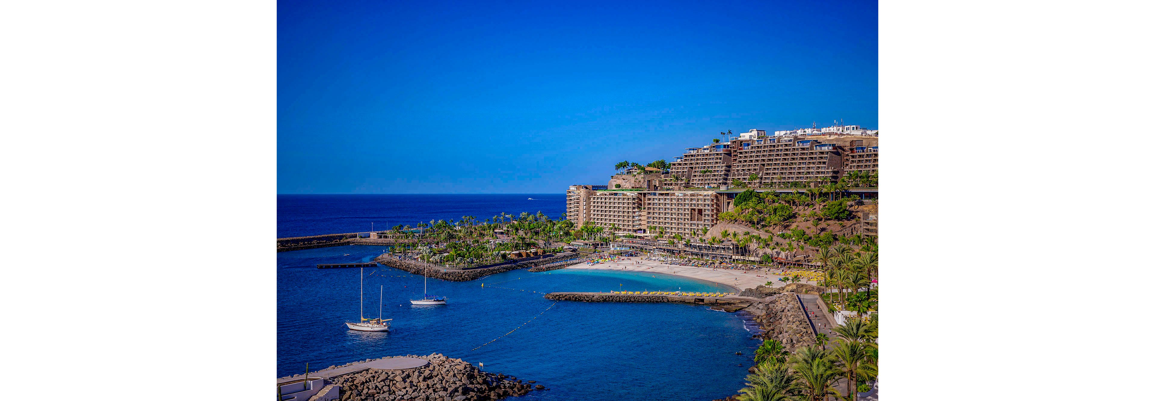 A large resort complex sits on a hillside overlooking a sandy beach and calm blue sea. Several sailboats are anchored near the shore, and palm trees line the waterfront, creating a tropical atmosphere. The sky is clear and vibrant, enhancing the inviting mood of the scene. The image captures a peaceful vacation destination with modern architecture and lush greenery. No visible text or numbers are present in the image.