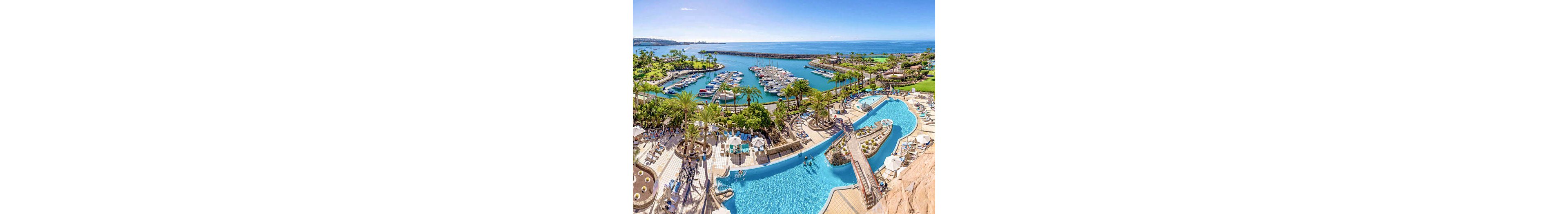 A large, curving resort swimming pool is set amidst palm trees and sun loungers, with guests enjoying the water and sunbathing. In the background, a vibrant marina filled with boats stretches out to a calm blue sea, bordered by lush greenery and a distant coastline. The scene is bright and inviting, with clear skies and a relaxed, vacation atmosphere. The pool area features multiple levels and bridges, adding visual interest to the setting.