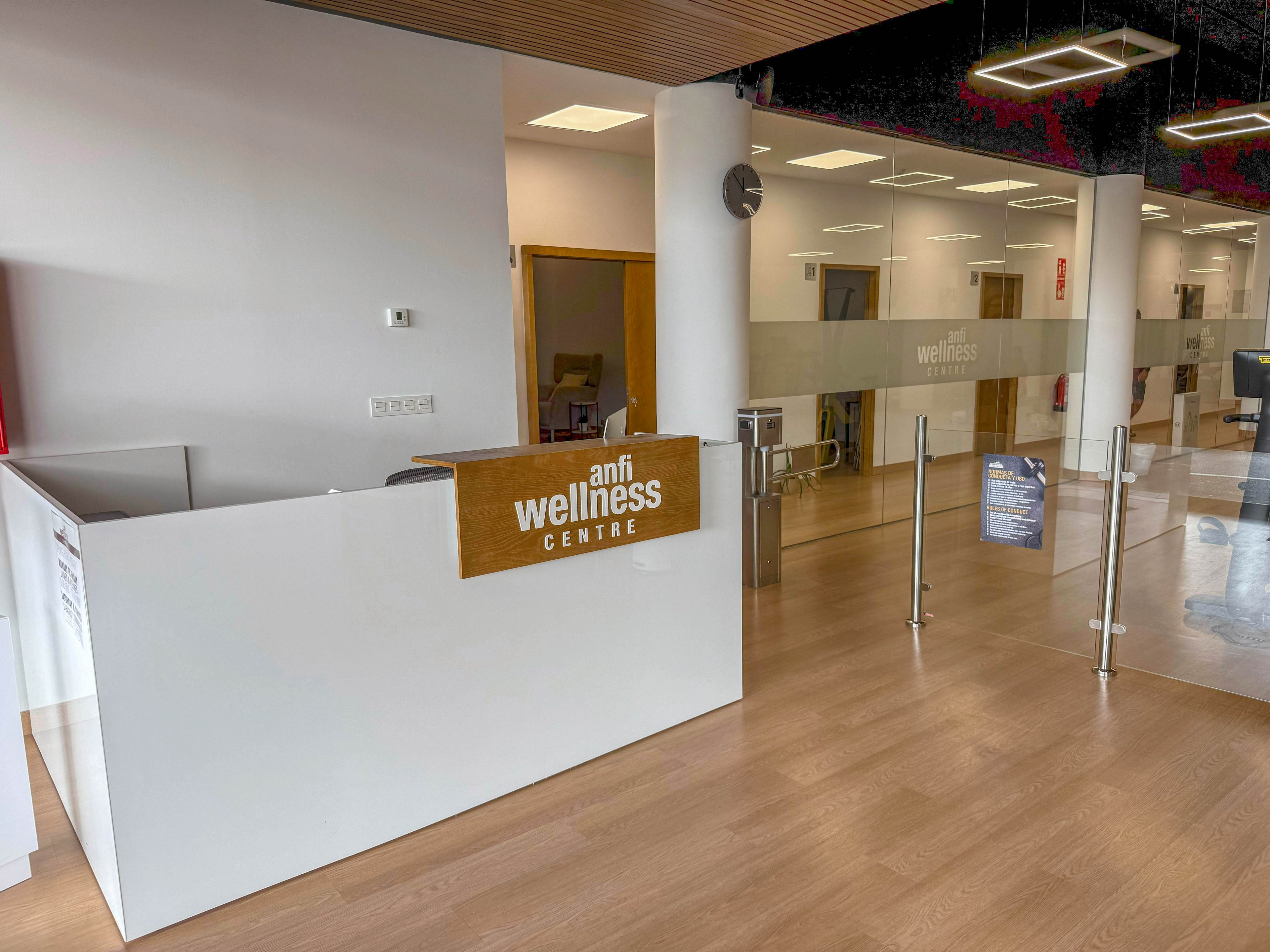 The image shows the reception area of the Anfi Wellness Centre, featuring a clean, modern design with a white desk and wood accents. Glass doors and walls create an open, inviting atmosphere, while the space is well-lit with geometric ceiling lights. The setting appears calm and professional, suitable for a wellness or health facility. The visible text includes 'anfi wellness CENTRE' on the reception desk.
