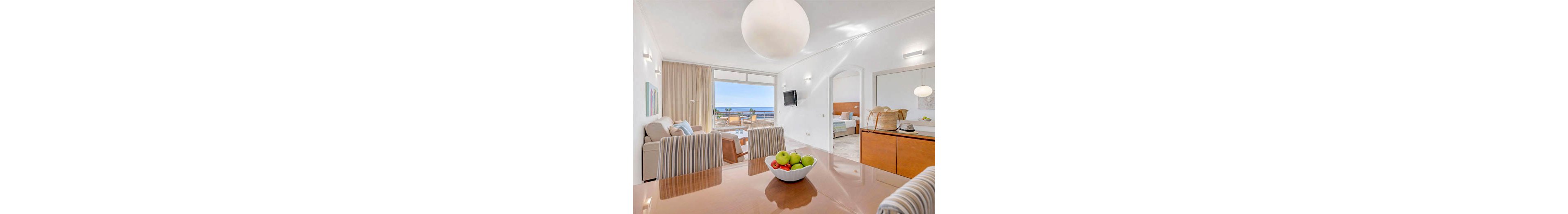 A spacious, sunlit apartment features a living area with striped chairs, a bowl of green apples on a polished dining table, and a cozy seating area. Large windows open to a balcony overlooking the ocean, creating an airy and inviting atmosphere. The adjoining bedroom is visible, and the decor is minimalist with soft neutral tones and contemporary lighting. No visible text or numbers are present in the image.