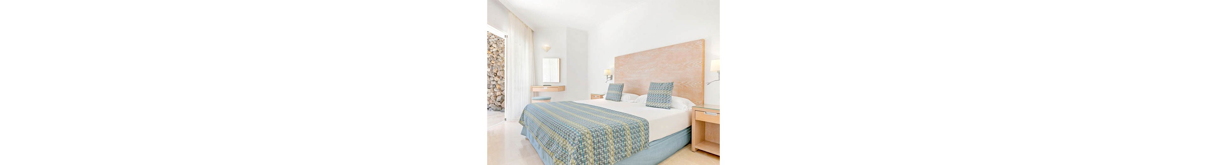 A spacious, sunlit hotel room features a large double bed with patterned blue and green bedding. The room is decorated in a minimalist style with light wood furniture, including a bedside table and a small desk with a mirror. Soft natural light enters through sheer curtains, and a stone wall is visible outside. The overall palette is light and airy, creating a calm and inviting atmosphere.