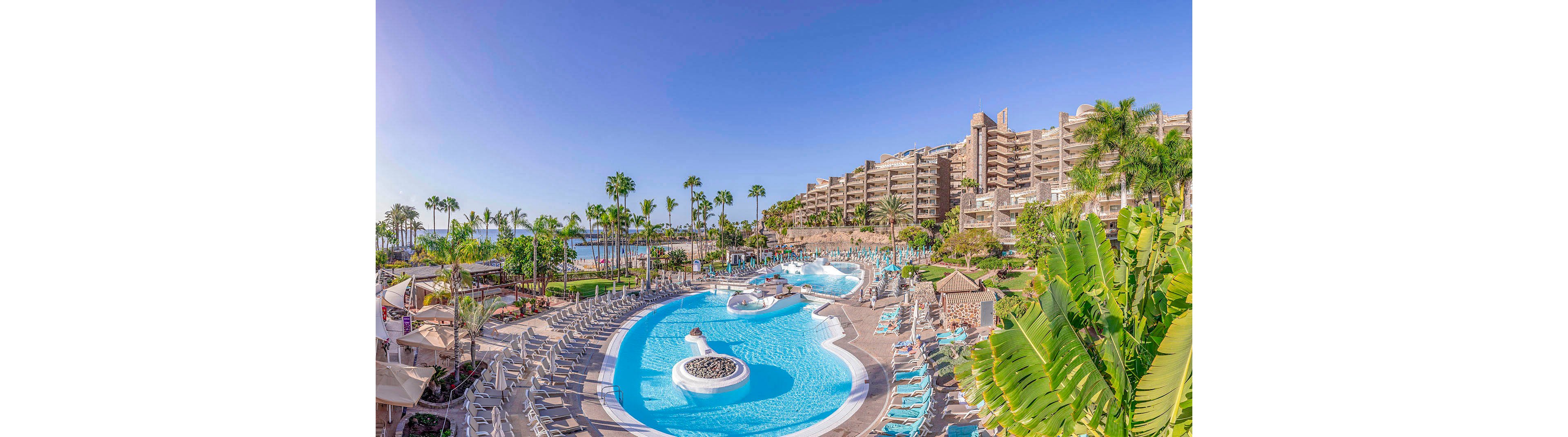 A large, curving outdoor swimming pool is surrounded by numerous lounge chairs and tropical palm trees. The resort buildings rise in the background, with balconies overlooking the pool and the distant ocean. The setting is bright and sunny, evoking a relaxed vacation atmosphere with clear blue skies and lush greenery. No visible text or numbers are present in the image.