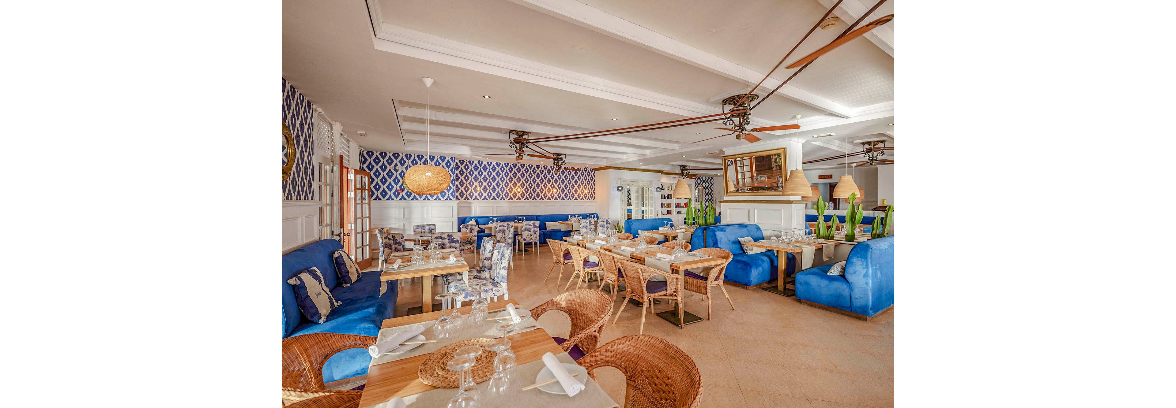 A spacious restaurant interior features a mix of wooden and wicker furniture, with plush blue sofas and patterned wallpaper. The setting is bright and airy, with natural light streaming in and contemporary ceiling fans overhead. Table settings include glassware and napkins, creating a welcoming dining atmosphere. The overall palette is dominated by blue, beige, and white tones, evoking a coastal, relaxed mood.