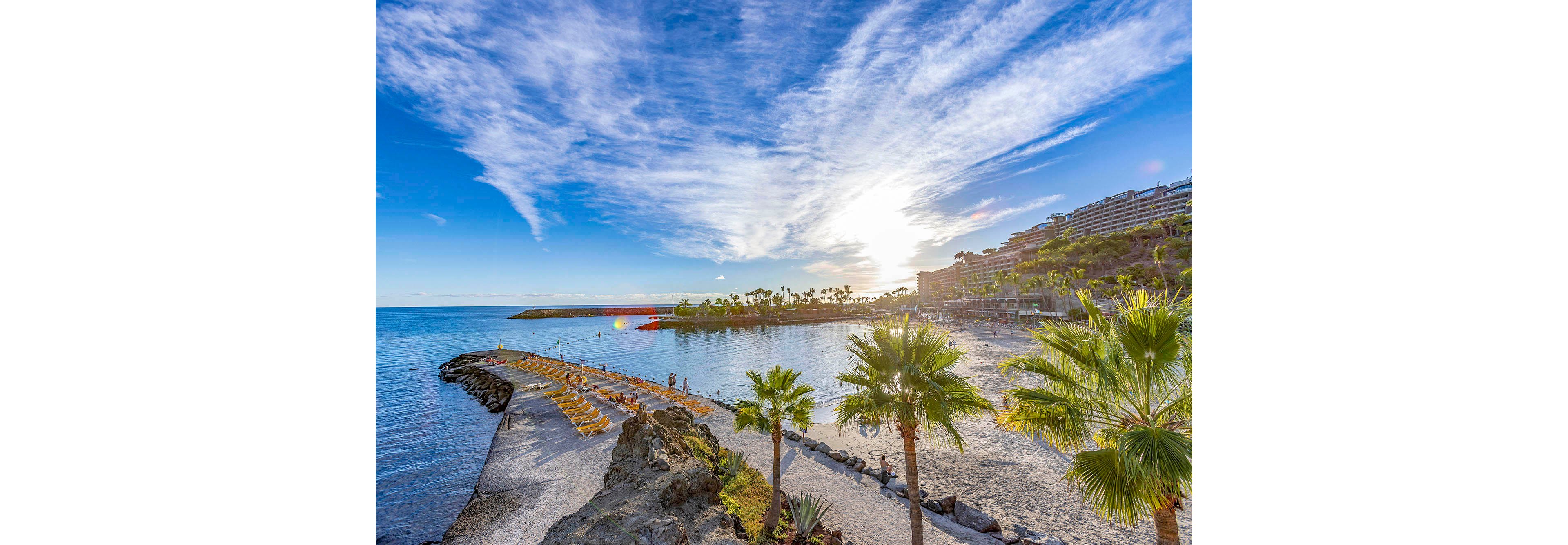 A vibrant tropical beach scene features tall palm trees and a calm shoreline under a bright blue sky. Sunlight reflects off the tranquil water, creating a serene and inviting atmosphere. Lounge chairs are arranged along the sandy beach, with a few people enjoying the peaceful setting. The background includes lush greenery and terraced buildings, enhancing the vacation vibe. The overall mood is relaxed and summery, with vivid colors and clear weather.