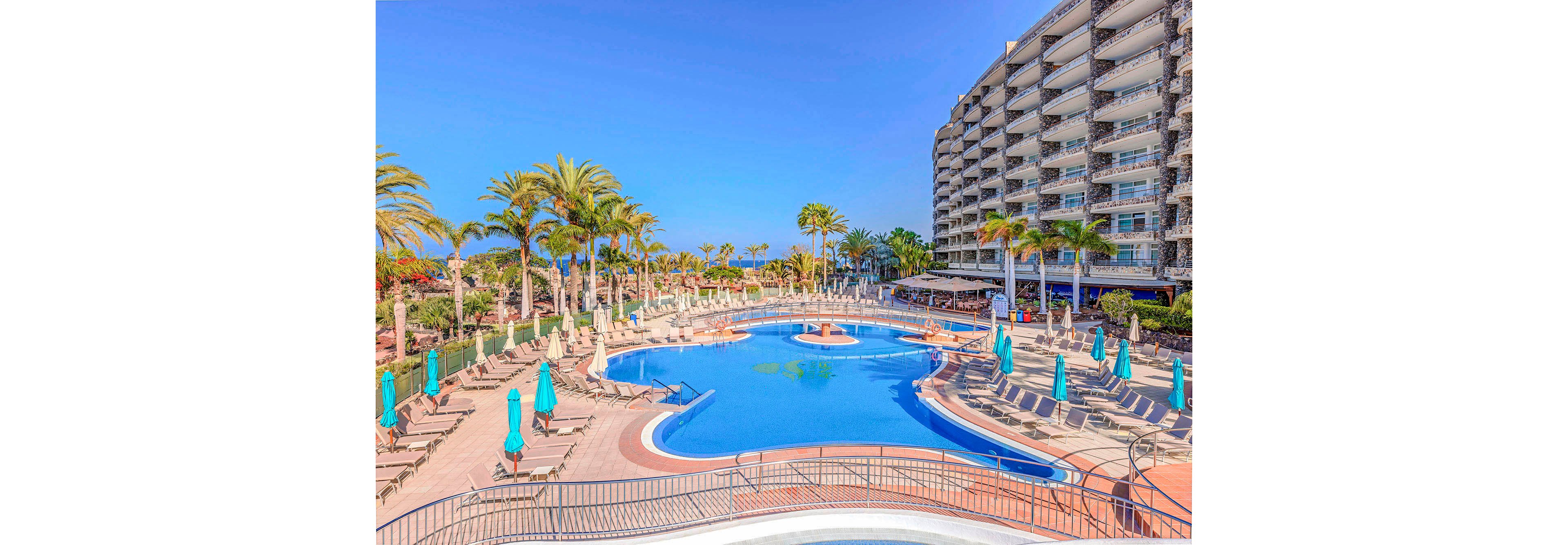 A large, inviting swimming pool is the central feature of this resort scene, surrounded by numerous sun loungers and turquoise umbrellas. Tall palm trees and lush greenery create a tropical atmosphere, while a multi-story hotel building stands prominently to the right. The clear blue sky and bright sunlight enhance the vibrant, relaxing mood of the setting. No visible text or numbers are present in the image.
