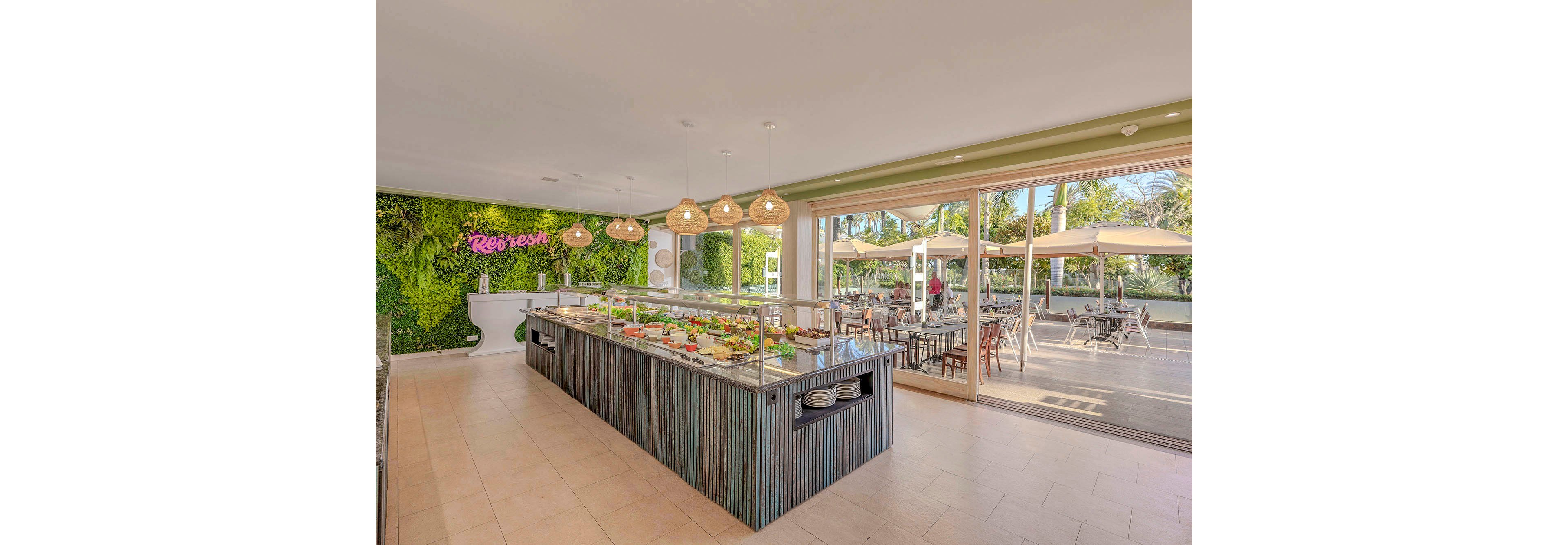 A contemporary salad bar is featured in a bright, airy dining area with large windows opening to an outdoor patio. The interior includes a green living wall with the word 'Refresh' in neon, pendant lighting, and a variety of fresh salads displayed under glass. Outside, tables and umbrellas provide shaded seating, creating a relaxed and inviting atmosphere. The overall palette is natural and fresh, emphasizing a healthy dining experience.