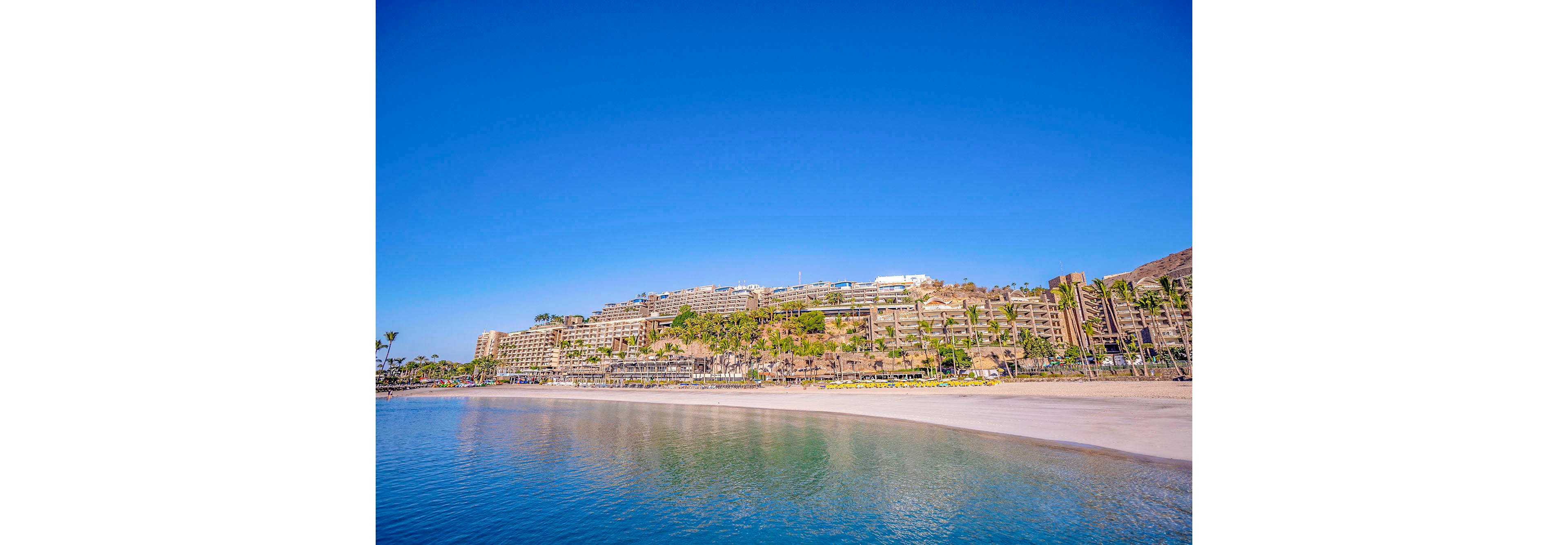 A series of large resort hotels are built into a hillside, overlooking a tranquil sandy beach and clear blue water. The scene is set under a vibrant blue sky, with palm trees and lush greenery visible around the hotels. The beach is wide and gently curves along the bay, creating a peaceful vacation atmosphere. No visible text or numbers are present in the image.