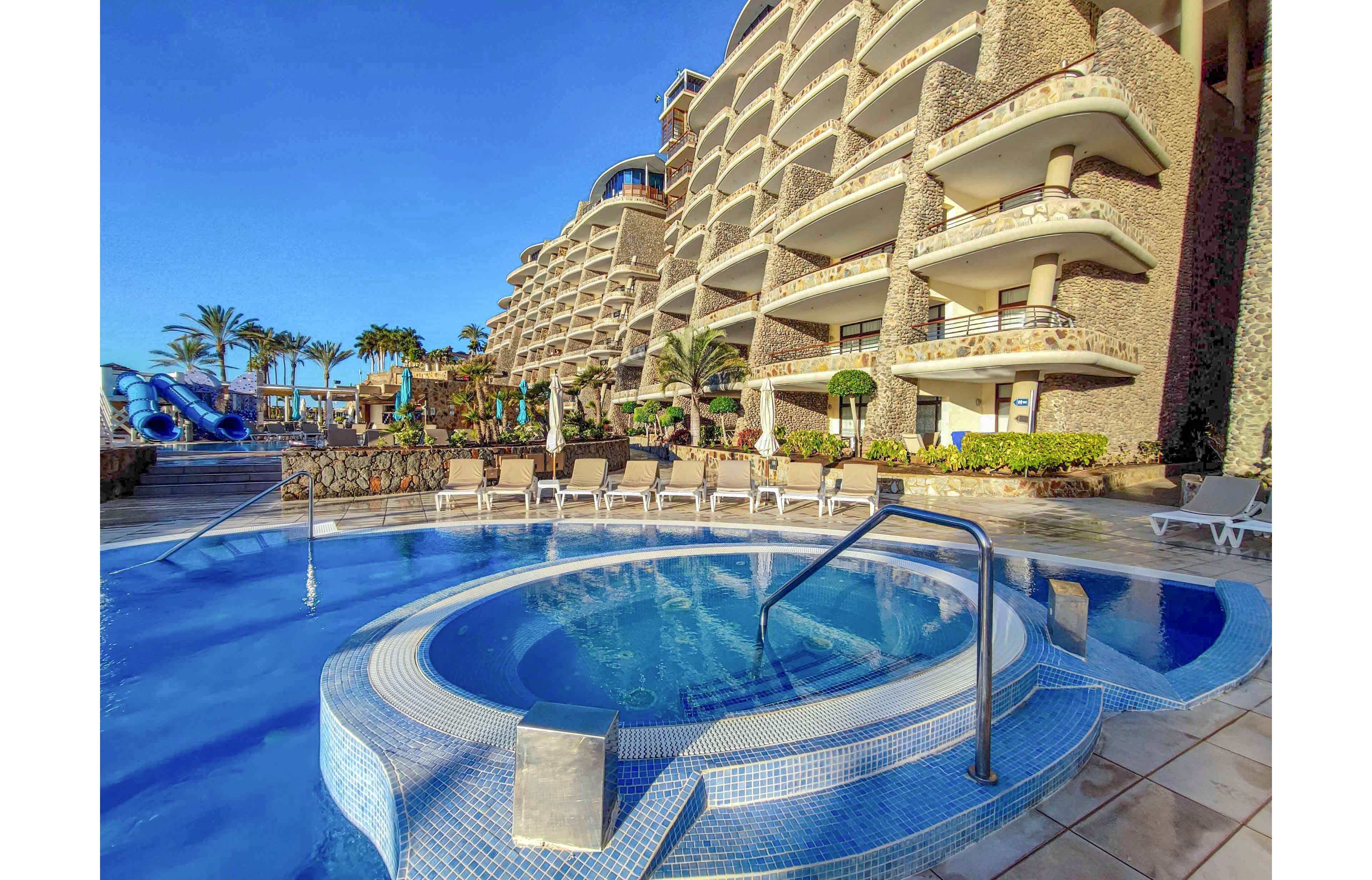 A circular hot tub is featured in the foreground, seamlessly connected to a large outdoor swimming pool. The setting is a modern resort with multi-story balconies, palm trees, and sun loungers under a clear blue sky. Water slides and lush landscaping add to the vacation atmosphere. The scene is brightly lit, evoking a relaxed and inviting mood. No visible text or numbers are present in the image.
