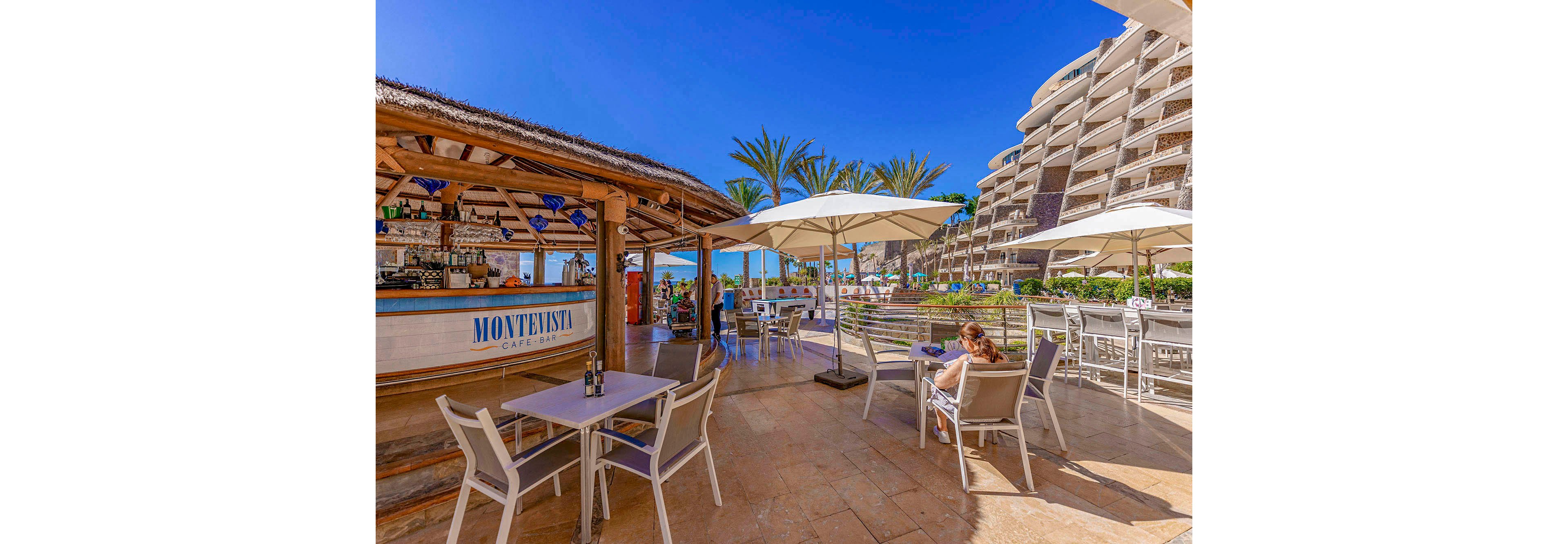 A vibrant outdoor café named 'Montenisa Café Bar' is set beside a modern resort building under a clear blue sky. The scene features shaded tables, white chairs, and large umbrellas, with guests relaxing and enjoying drinks. Palm trees and the ocean are visible in the background, creating a relaxed, sunny atmosphere. The setting is bright and inviting, with a Mediterranean coastal vibe.