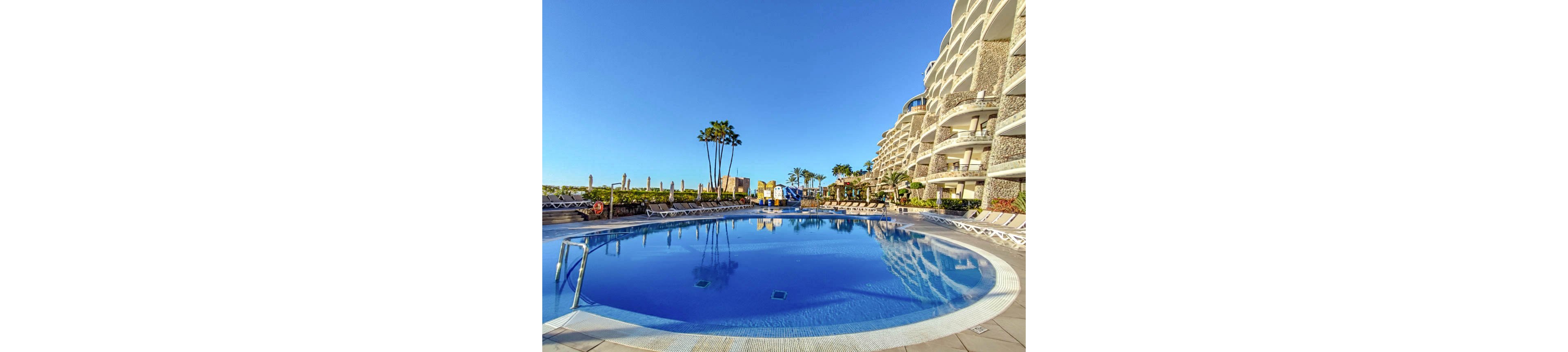 A large, circular outdoor swimming pool is the main focus, surrounded by numerous sun loungers and umbrellas. The pool is adjacent to a modern, multi-story hotel with curved balconies, and a few tall palm trees add a tropical touch. The sky is clear and blue, creating a bright and inviting atmosphere, with no people visible in the scene.