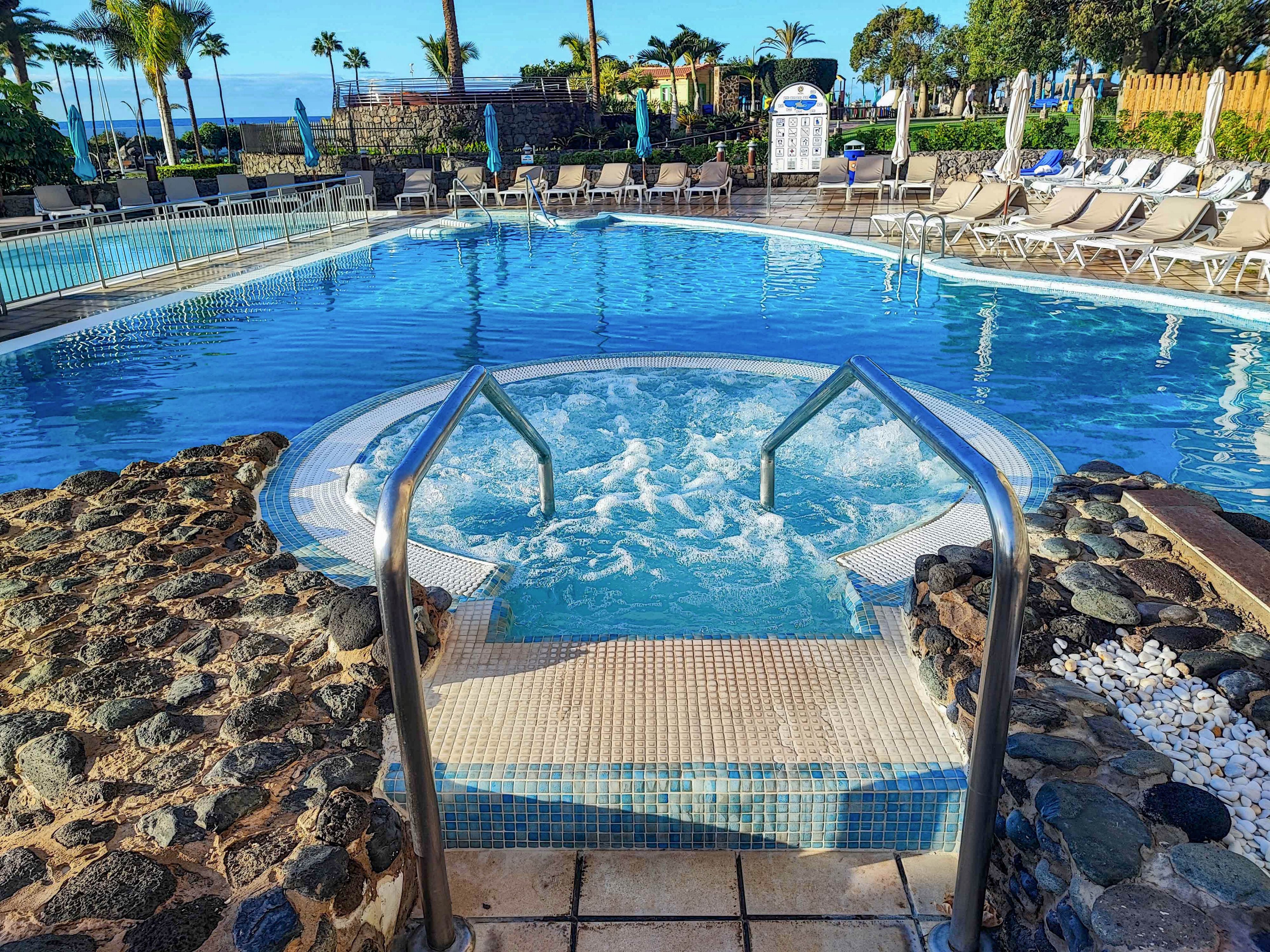 A bubbling hot tub spa is set in the foreground, leading into a large, clear blue outdoor swimming pool. The scene is surrounded by lounge chairs, palm trees, and umbrellas, evoking a relaxing vacation atmosphere. The setting is bright and sunny, with a tropical resort vibe and no visible people. The pool area is bordered by stonework and landscaped greenery.