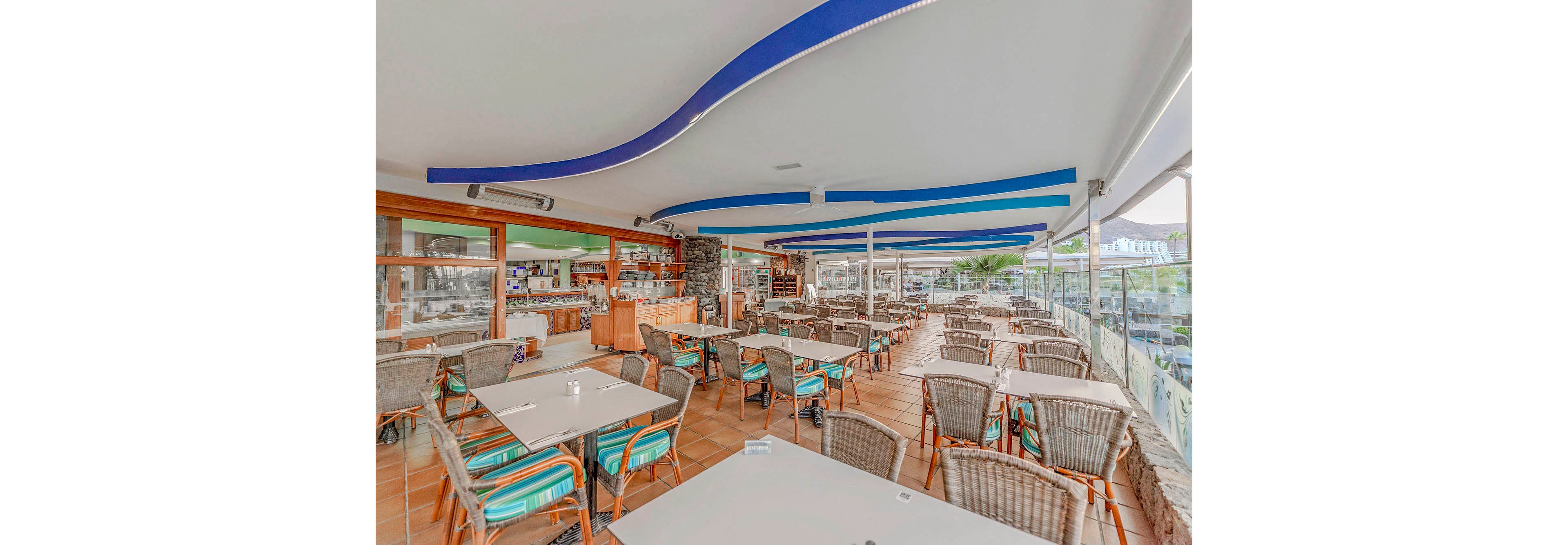 A large, open-air restaurant features numerous wicker chairs and tables arranged neatly under a covered patio. The setting is bright and airy, with blue wave-like ceiling accents and glass walls providing a view of the outdoors. The atmosphere is casual and inviting, with a mix of natural and modern elements. No people are present, highlighting the clean and organized dining area. The color palette includes neutral tones with pops of blue and green.